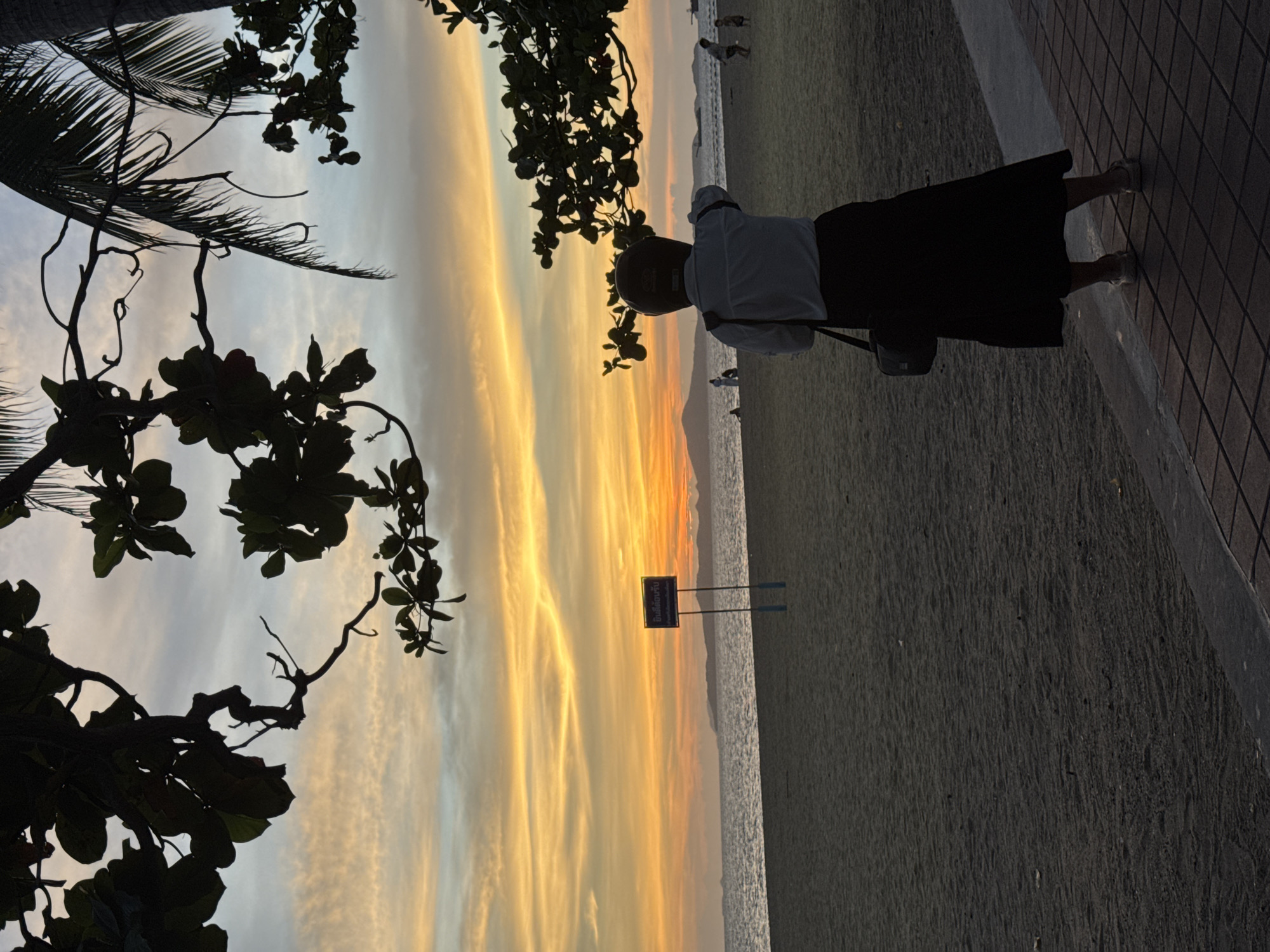 A woman in a light-colored shirt and dark skirt stands on a tiled walkway along a beach in Pattaya City, Thailand. She is facing away from the camera, towards the sea. The beach is sandy, and the water stretches to the horizon. A sunset paints the sky in shades of yellow and orange, with clouds streaked across the expanse. Silhouetted trees frame the scene on the left side of the image. A sign is visible near the water's edge, and other people are scattered along the beach.