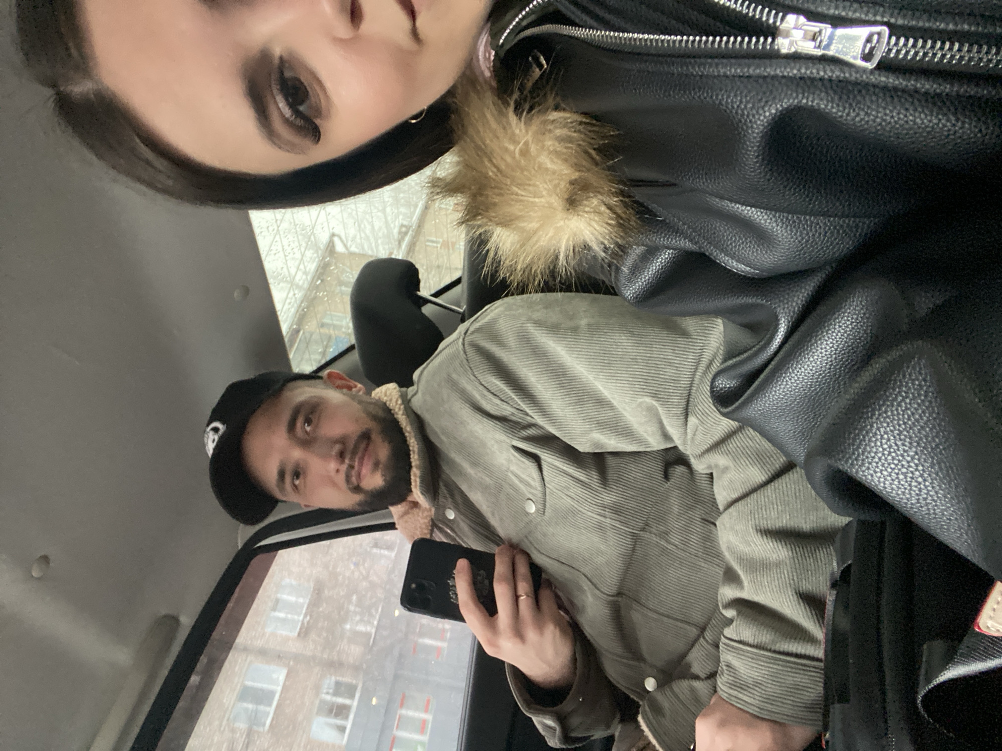 A selfie-style photograph captures two individuals inside a vehicle. A woman with dark hair and prominent eye makeup occupies the upper portion of the frame, partially obscured. The second person, a man with a beard wearing a corduroy jacket, gazes toward the camera while holding a black smartphone. He is wearing a baseball cap. The interior of the vehicle is visible, with a car seat headrest and a window behind the man. Through the window, a multi-story building is visible, indicative of an urban environment. Based on the provided location, Chaikovsky, Russia, this suggests the photo was taken within that city. No specific street name or building is identifiable.