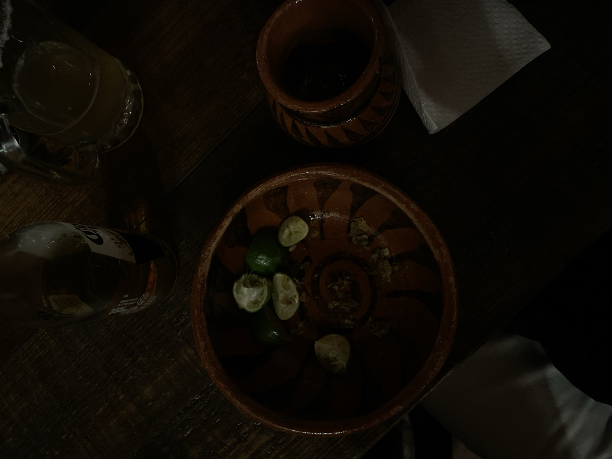 The image displays a dimly lit tabletop scene, likely inside a bar or restaurant in Puebla, Mexico, during nighttime. The surface appears to be a dark wooden table. In the upper left, a clear glass beer mug, partially filled with a light-colored beverage, is visible. Below it, a clear glass beer bottle, possibly a Corona, lies on its side, though most of its label text is obscured by darkness and reflection.
In the top center, a traditional reddish-brown terracotta pot, adorned with geometric patterns, is present, holding an indiscernible dark liquid. The most prominent item in the center is a round, shallow terracotta bowl. Inside this bowl are several green lime wedges, some whole and some already squeezed, accompanied by small, irregular brown pieces that might be dried spices, nuts, or chili flakes. A folded white paper napkin rests on the table in the upper right corner. The traditional terracotta ware and the presence of limes suggest a typical Mexican setting, indicating a casual social gathering or meal. The overall mood is relaxed and informal, characteristic of a night out.
