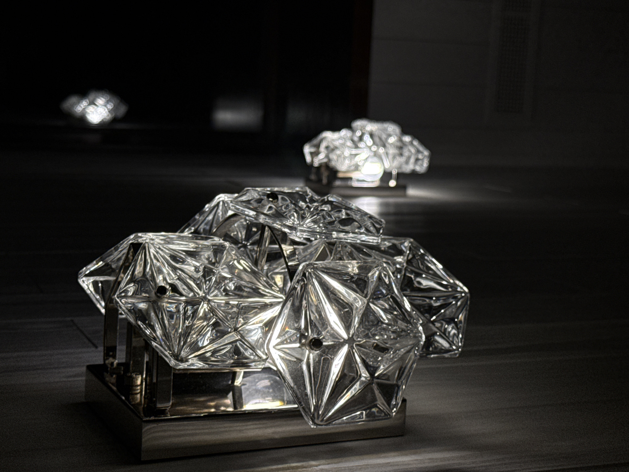 The image shows a close-up view of a modern glass wall sconce. The sconce features multiple faceted crystal or glass elements clustered together, creating a geometric and sculptural design. The material is highly reflective, catching the light and casting intricate patterns. The sconce is mounted on a brushed metal fixture against a dark, textured wall. In the background, out of focus, another similar sconce is visible, emitting a warm light. The scene appears to be an interior setting, possibly a hotel or a high-end residence. The location within the city of Urlu-Aspak, Russia, is not discernible from the visual information provided.