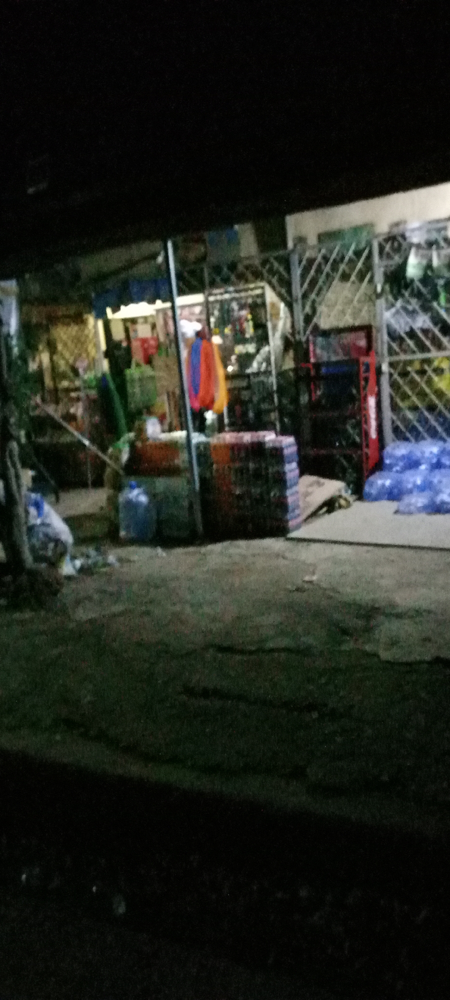 The image depicts a nighttime scene in Jos, Nigeria, showing the exterior of a small shop or store. The foreground consists of a dark, grassy or dirt area, possibly the roadside or sidewalk. The shop's entrance is partially visible under a roof, with shelves and displays of various merchandise inside. Outside the shop, there are stacks of crates or containers, a large blue water container, and piles of plastic bags. To the right, a red Coca-Cola branded display rack and a metal security gate are visible. The lighting is dim, with illumination coming from inside the shop, casting shadows on the foreground. The overall impression is that of a small, local business operating in a low-light environment. There are no people visible in the image.