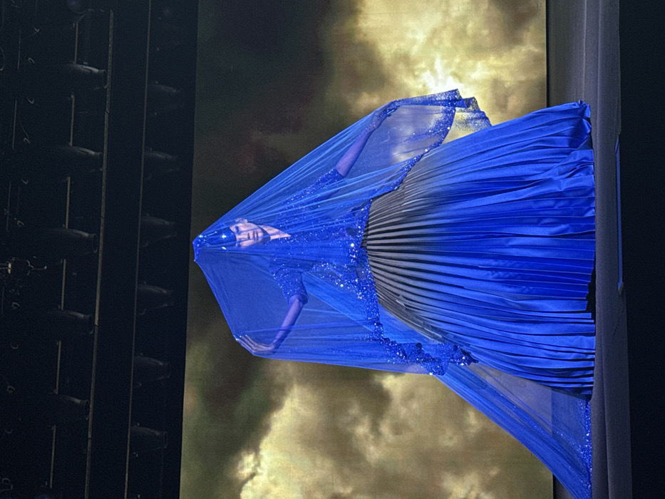 A figure draped in shimmering blue fabric stands against a backdrop of swirling gold and brown hues. A matching blue veil, also adorned with glittering accents, partially obscures the face. The figure's arms are visible beneath the veil. The lower portion of the figure's dress is a pleated material. To the left, a dark structure with repeating rectangular shapes appears, contrasting with the light and color of the central figure.