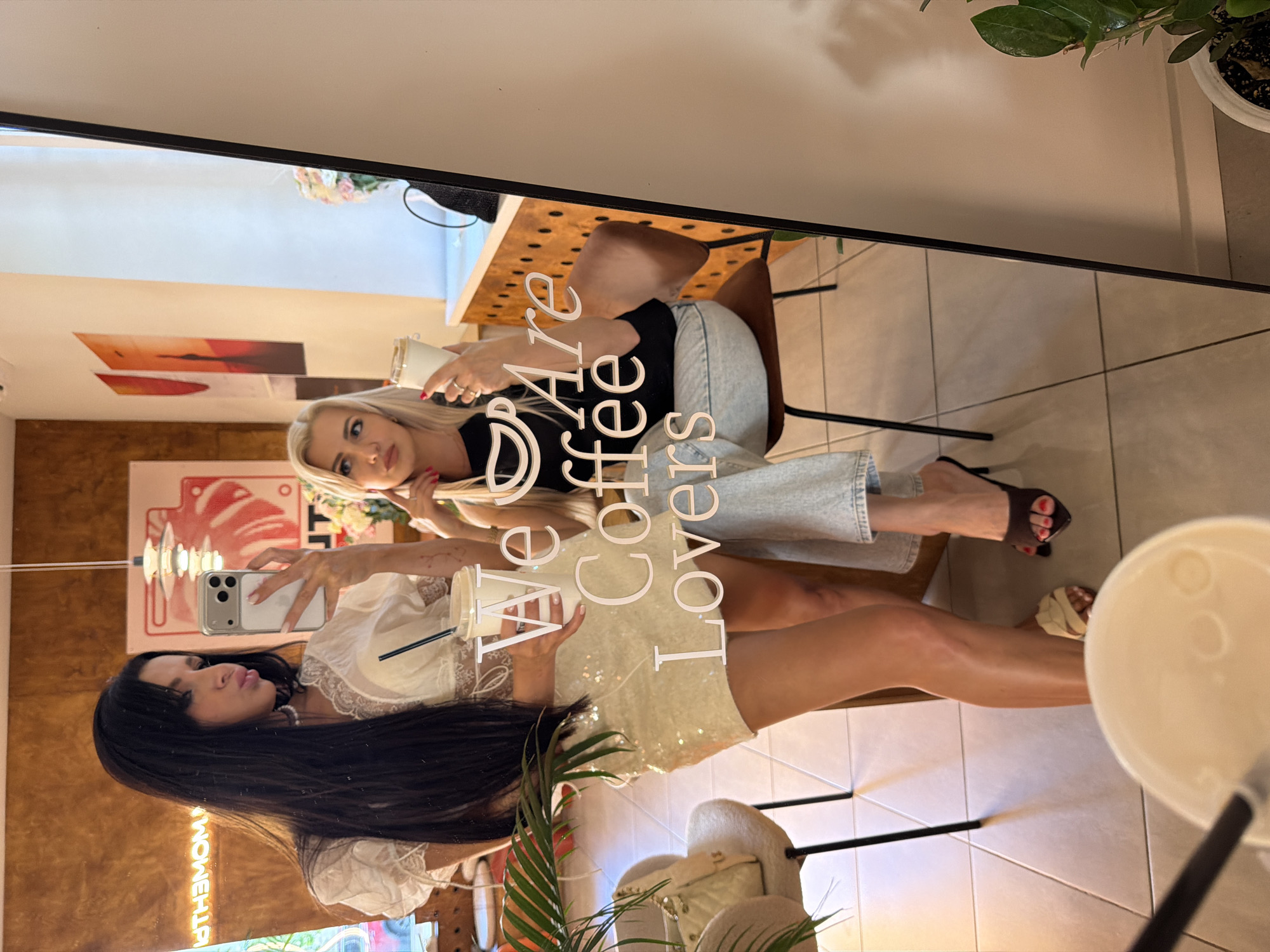 Two women are posing for a selfie in a cafe. The woman on the left has long dark hair and is wearing a white lace top and a sequined skirt. She is holding a smartphone and a white cup with a straw. The woman on the right has blonde hair and is wearing a black t-shirt and jeans. She is holding a white cup. The cafe has a warm, rustic interior with wooden accents and art on the walls. The text "We Are Coffee Lovers" is visible on a mirror. The scene is set in Novaya Usman', Russia, likely within a cafe called "The Moment."