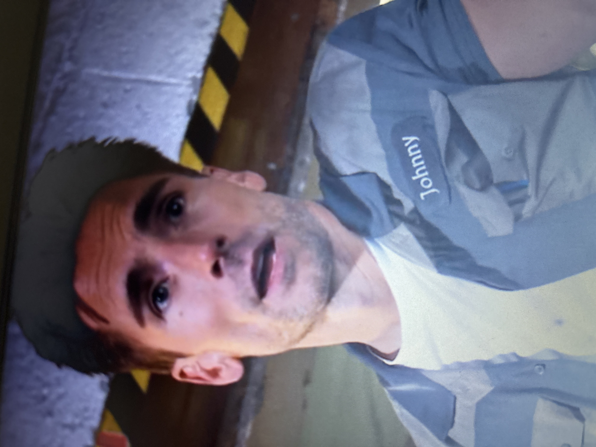 A man with light brown hair and eyes, wearing a light blue work shirt with the name "Johnny" embroidered on it, is looking upward with a slightly open mouth. He is standing near a wall marked with black and yellow stripes. The location is unidentified.