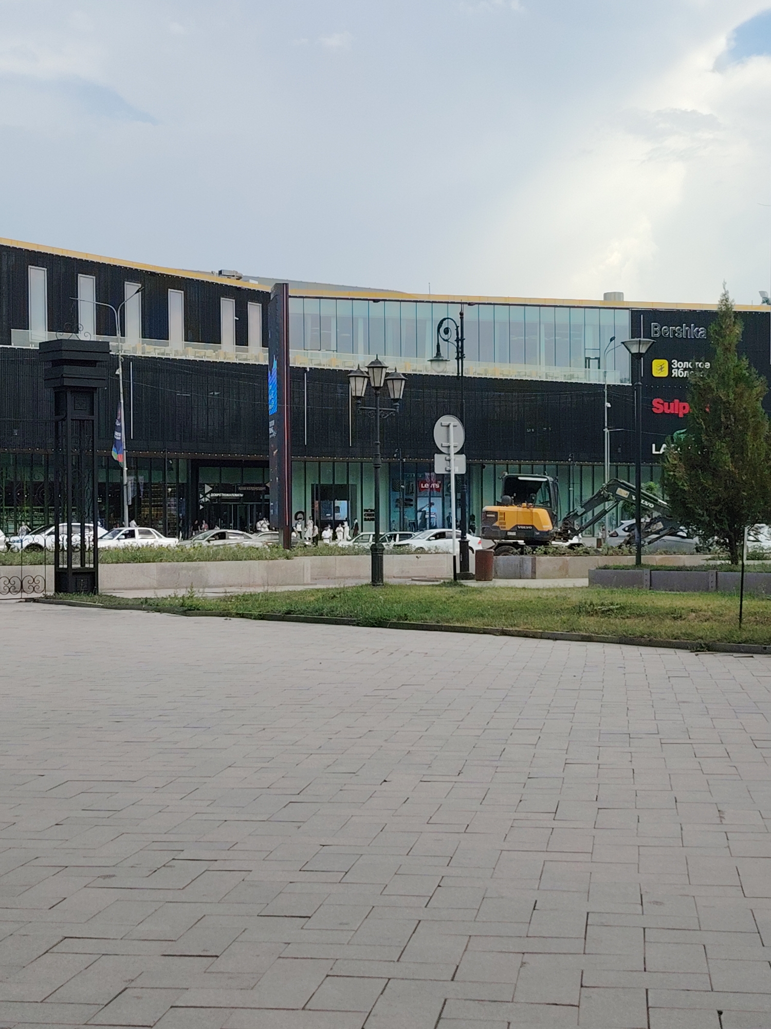 The image shows the exterior of a modern shopping mall in Shymkent, Kazakhstan, under a cloudy sky. In the foreground, a paved area with a repeating pattern of rectangular stones leads towards the mall. The mall features a dark, textured facade with large glass windows and a yellow trim along the roofline. Several stores are visible through the windows, including "Bershka" and "Sulpak." An excavator is positioned in front of the mall, suggesting ongoing construction or landscaping work. Several cars are parked in front of the mall, and a few people can be seen walking around the entrance. The overall scene depicts a commercial area in a city, with elements of both retail activity and urban development.