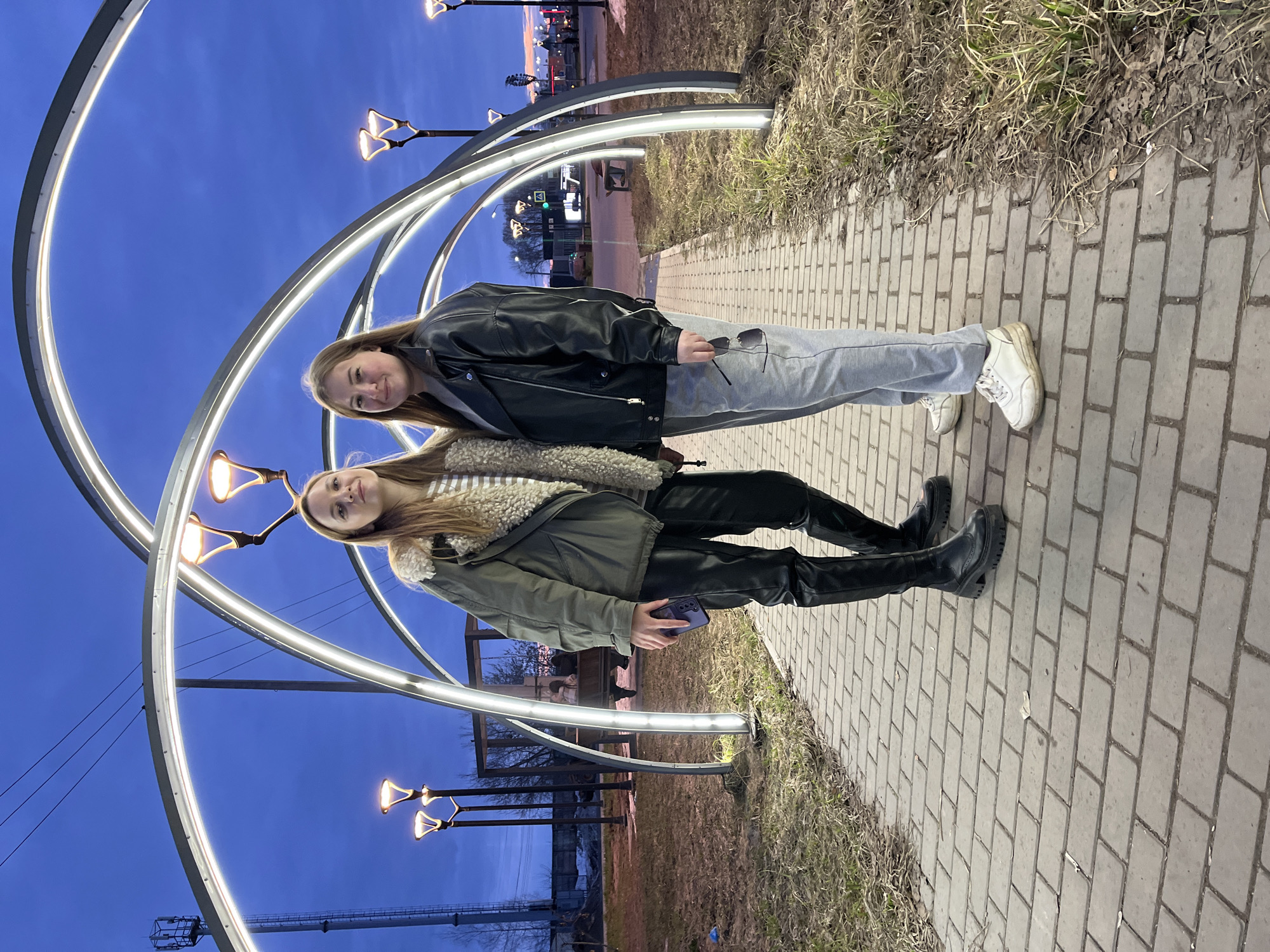 Two young women are standing side-by-side on a brick-paved walkway under an illuminated metal tunnel structure. The location is likely a public space in Chaikovsky, Russia, given the user's request. The woman on the right wears a black leather jacket, grey sweatpants, and white sneakers, holding sunglasses in her hand. The woman on the left wears a green jacket with a furry collar, black leather pants, and black boots, holding a smartphone in her hand. The background includes other light fixtures, a grassy area, and glimpses of urban structures and a twilight sky.