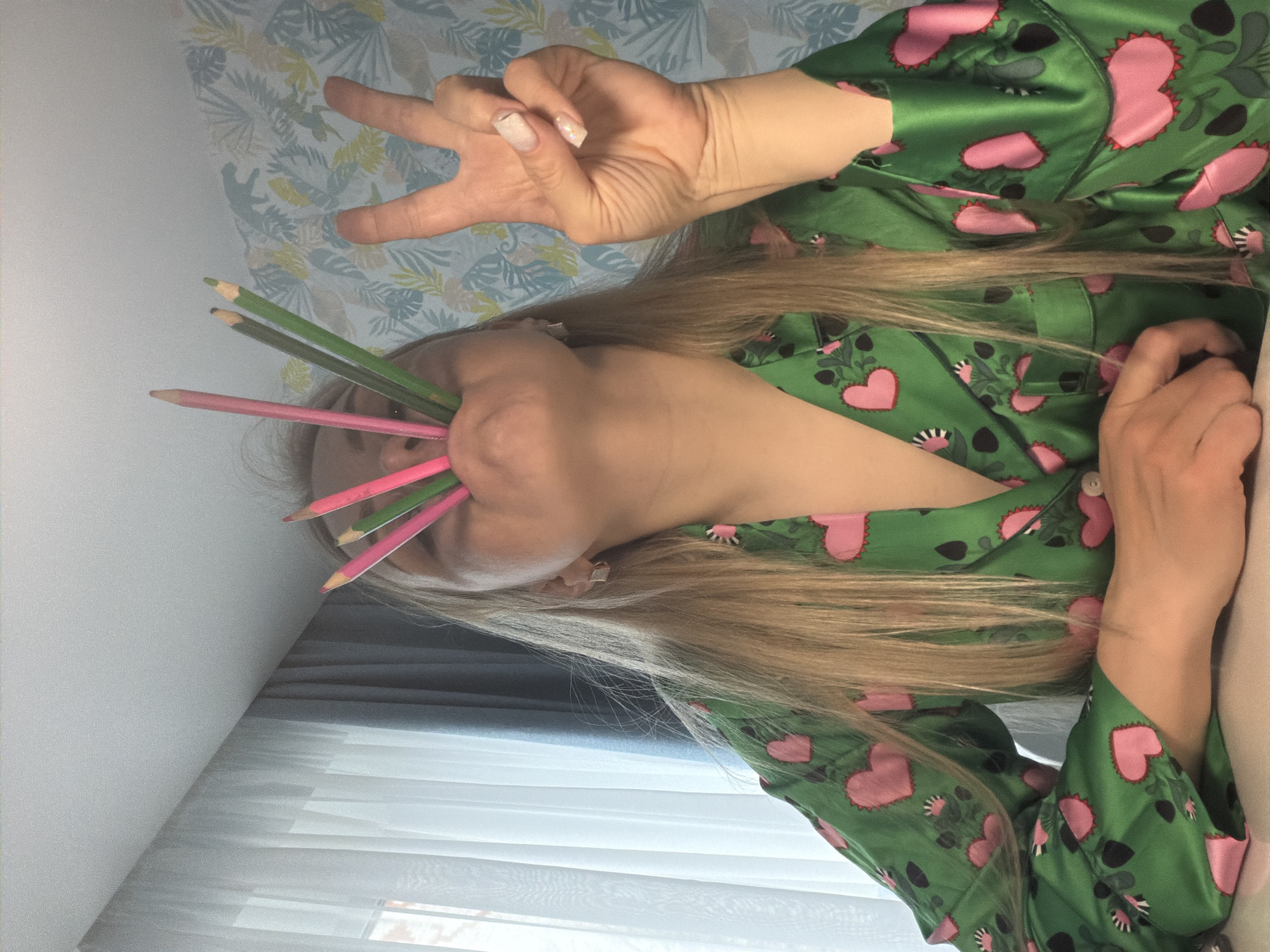 A close-up, rotated portrait-style image captures a person, likely female, with long blonde hair, posing in an indoor setting. The individual is wearing a vibrant green pajama top patterned with numerous pink hearts and smaller dark green floral designs. Several colored pencils, predominantly green and pink, are playfully held in the person's mouth, partially obscuring their face.
The person's right hand, positioned in the upper part of the frame, is raised and forms a peace sign (V-sign) with two fingers. The fingernails of this hand display a light-colored, possibly glittery, manicure. The left hand is partially visible, resting towards the bottom right of the frame.
The background features a light-colored wall, the upper half of which is adorned with a decorative wallpaper showcasing light blue, green, and yellow botanical motifs. Below the wallpapered section, a window covered by white horizontal blinds or curtains is visible, allowing diffuse daylight into the room. The overall lighting suggests it is daytime. The playful pose, incorporating the pencils and peace sign, conveys a lighthearted and relaxed mood. No text is visible in the image. The setting appears to be a comfortable domestic space, likely in Votkinsk, Russia.