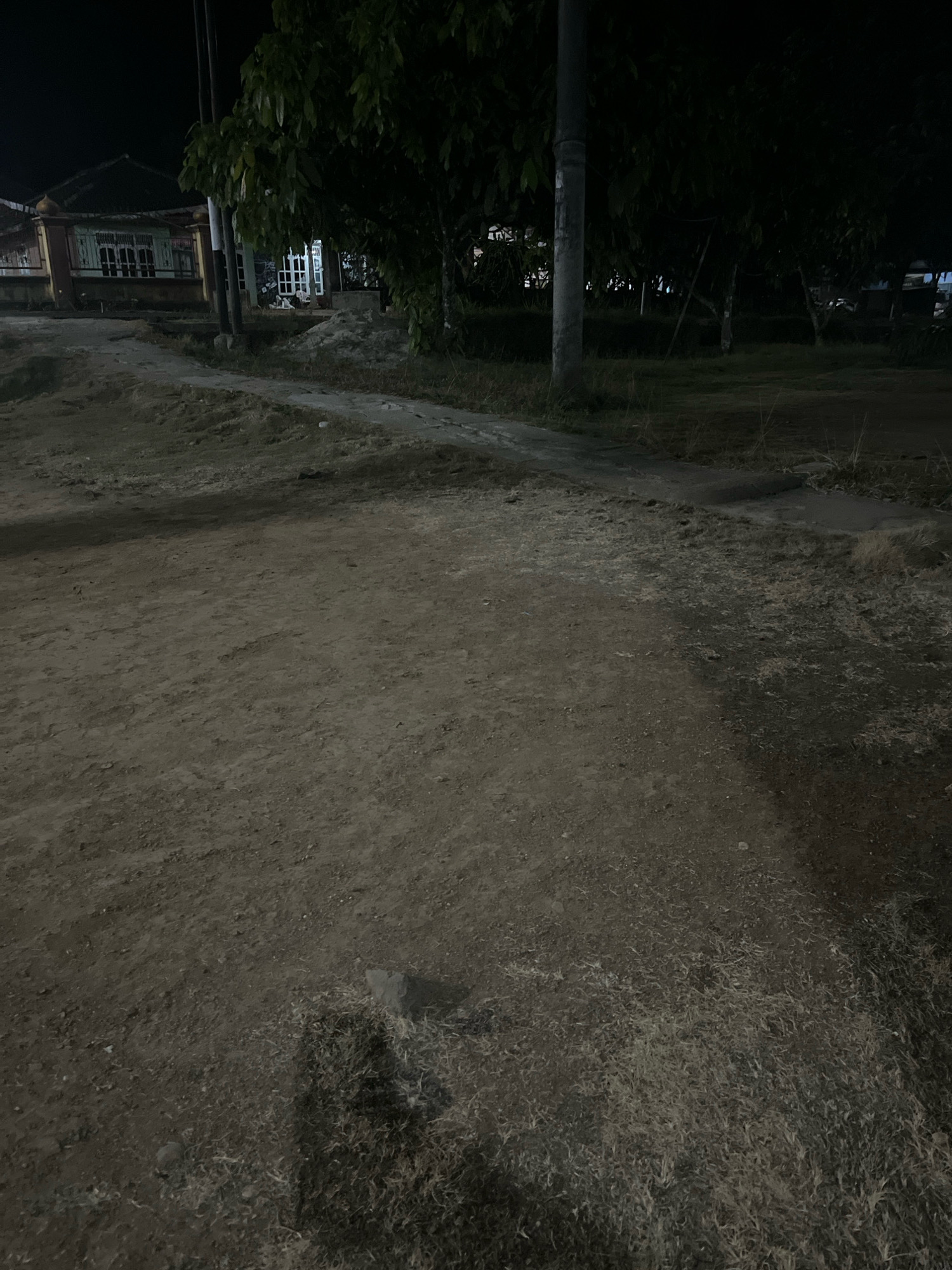 The image depicts a dark, outdoor scene, likely taken at night, showing a dirt path winding through a residential area. In the background, a house with a dark roof, white-framed windows, and visible interior lighting is partially obscured by trees and foliage. A vertical pole, possibly a utility pole, stands prominently in the mid-ground. The ground in the foreground consists of dry, uneven dirt with patches of dry grass and small stones. The lighting conditions suggest artificial illumination from the house and potentially streetlights, casting shadows across the scene. The overall impression is one of a quiet, possibly rural or suburban setting at night. The location within the city of Kebumen, Indonesia, is suggested by the architectural style of the house and the general environmental characteristics which align with tropical or subtropical regions.