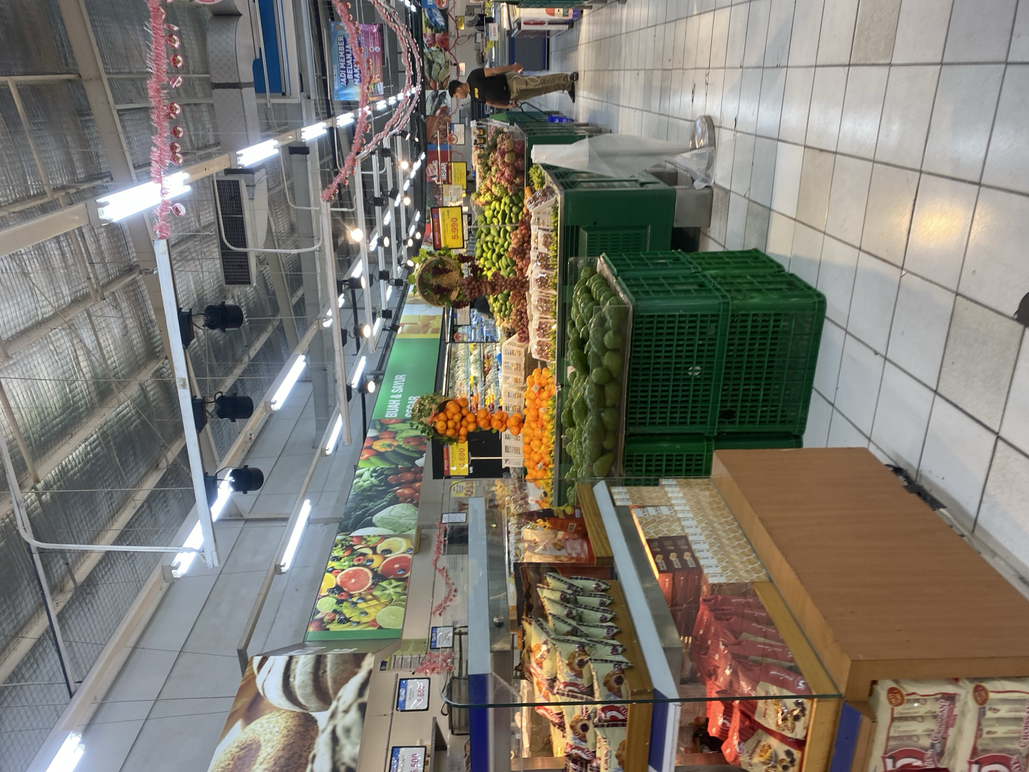 This is a photograph of the interior of a supermarket in Banjarbaru, Indonesia. The scene depicts a produce section with a variety of fruits and vegetables displayed. In the foreground, a refrigerated display case contains packaged snacks, and to the right, a man wearing a black t-shirt and khaki pants stands near a display of grapes and avocados. Green plastic crates filled with produce are visible throughout the aisle. Overhead, fluorescent lights illuminate the store, and promotional banners hang from the ceiling. The floor is tiled with a light-colored pattern. The overall impression is a well-stocked grocery store with an emphasis on fresh produce.