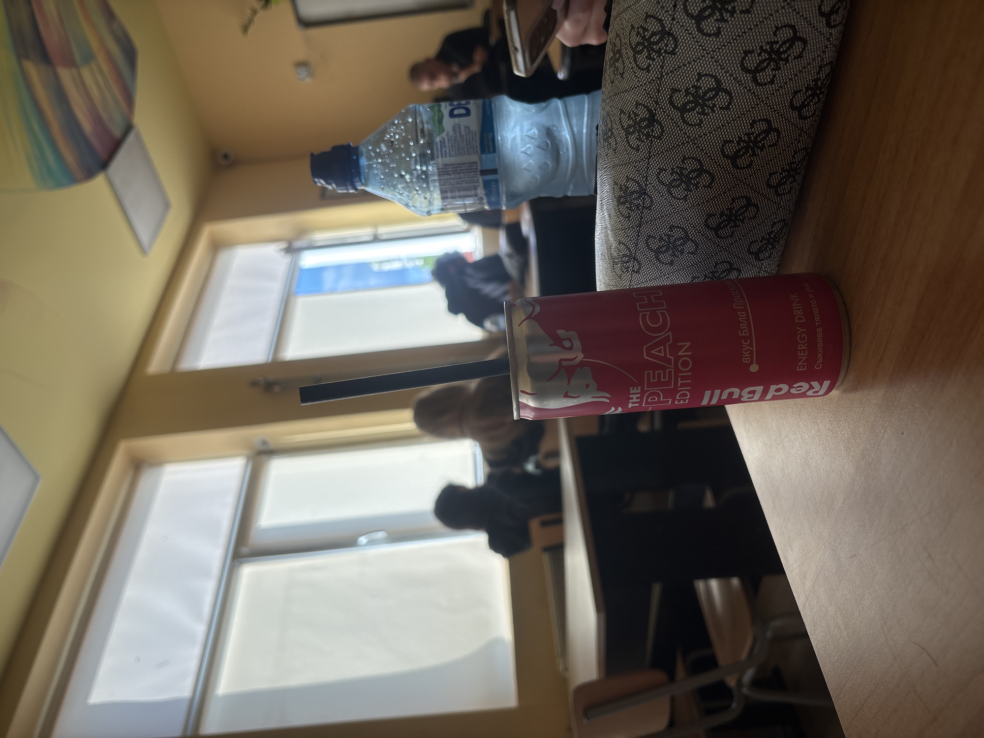 The image shows a desk in a room, possibly a classroom, with a Red Bull Peach Edition can, a water bottle labeled "DE," and a black and white patterned bag. A black straw is extending from the top of the can. There are blurred figures of people in the background, sitting at desks. The windows show bright daylight, suggesting daytime. The location is likely in Sofia, Bulgaria, given the Bulgarian text on the Red Bull can. The primary subjects are the drink and bottle on the desk.