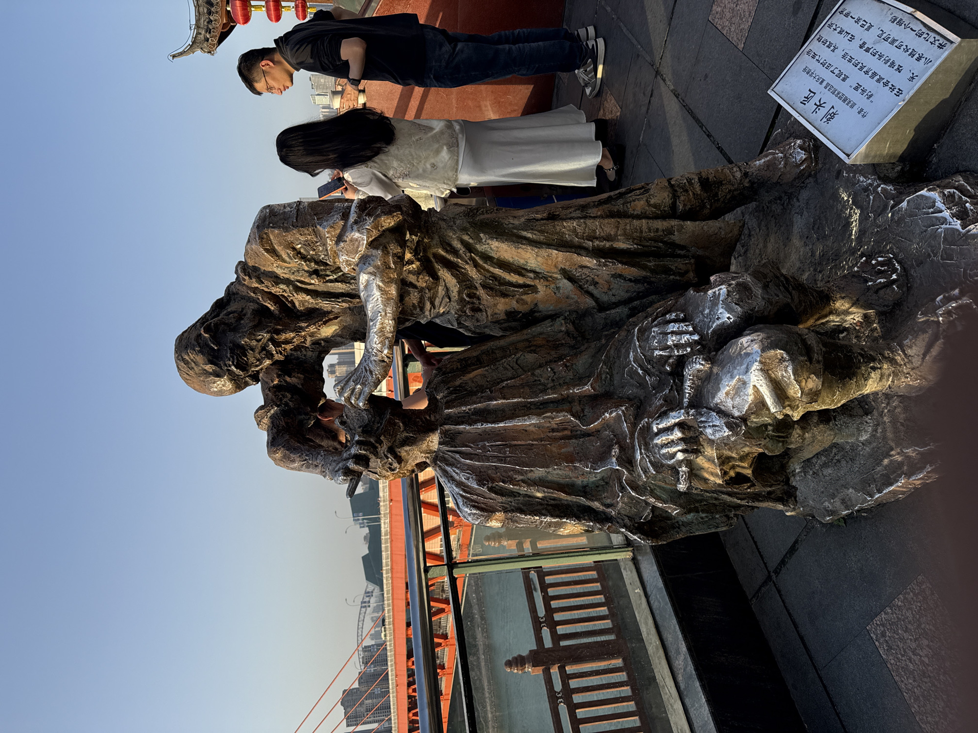 A photo shows a bronze statue of two figures, presumably a "剃头匠" (Tìtóu jiàng - barber) and customer in the city of Yuzhong District, China. A man in a black t-shirt and dark jeans, and a woman wearing a long white dress with a white jacket, stand behind it. A sign describing the artist and the statue is to the right. Red girders appear on a nearby bridge. The sky is clear blue.