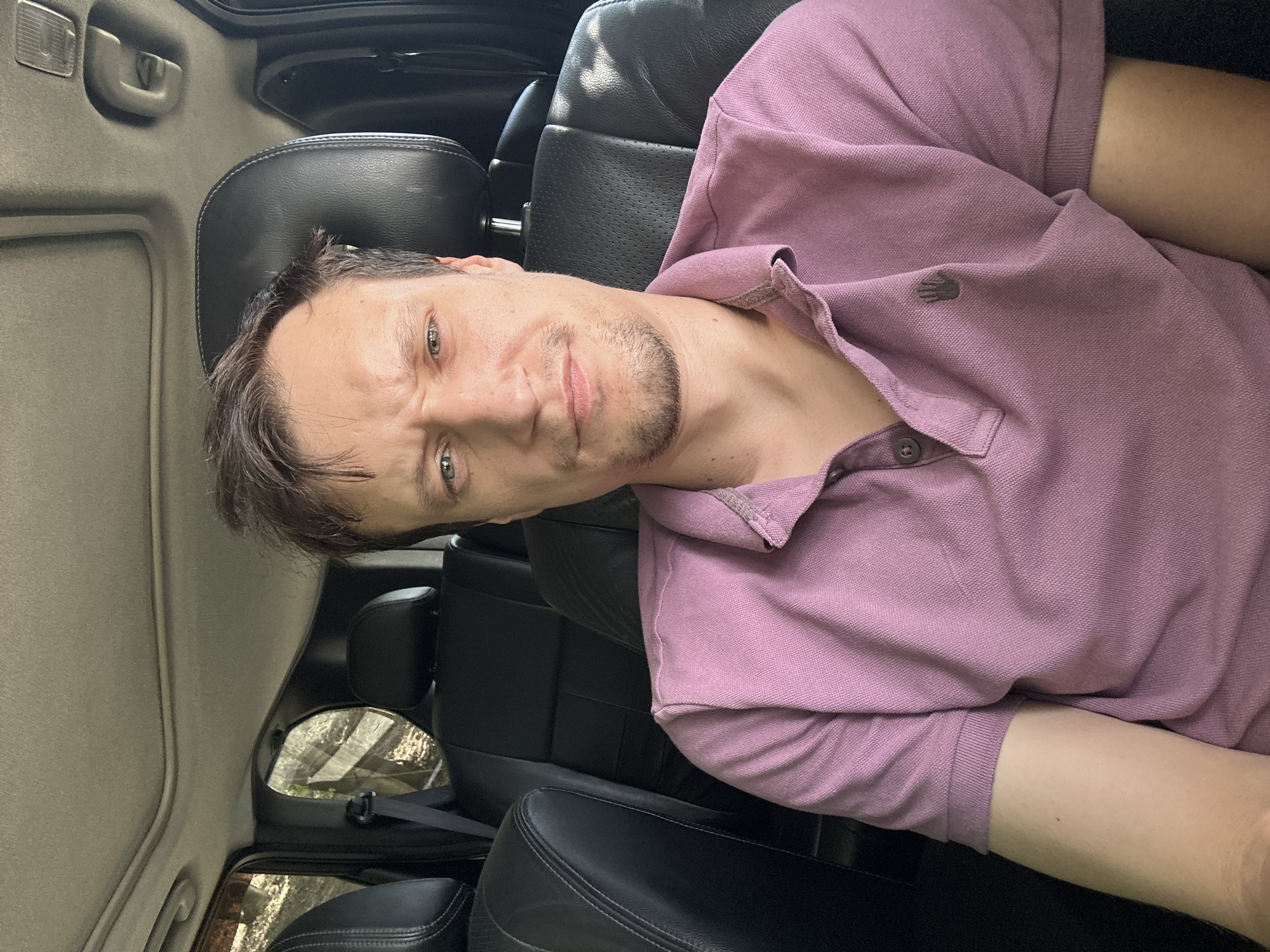 The image shows a man sitting in the back seat of a vehicle. He is looking directly at the camera with a neutral expression. The man has short, brown hair and is wearing a pink polo shirt. The interior of the vehicle is dark, with leather seats visible. The background outside the vehicle appears to be a wooded area, suggesting the car may be parked on a street or in a rural location. There is a sunroof open above the man. There is no indication of the city of Makiv, Ukraine within the image.