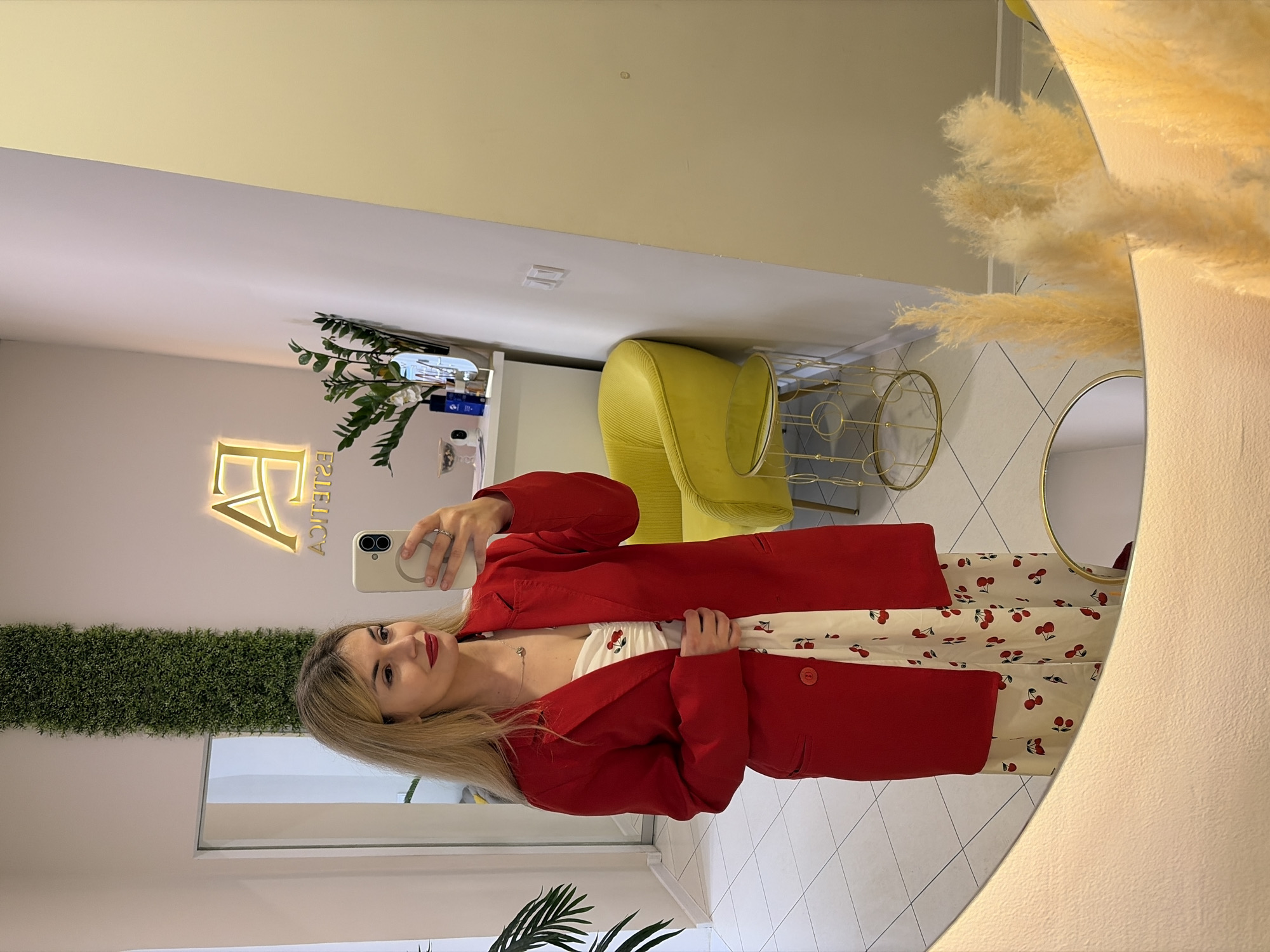 A young woman with long blonde hair, wearing a red blazer over a cherry-patterned top, is taking a selfie in a stylish interior. She is holding a phone in her right hand. The scene is reflected in a round mirror decorated with pampas grass. In the background, there's a yellow chair and a gold-colored side table. On the wall is a sign with the text "FA ESTETICA". Considering the aesthetic and the sign, the location is likely a beauty salon or clinic, possibly in Budva, Montenegro. The woman is the main subject, engaged in the action of taking a selfie within this stylish interior. The reflection in the mirror indicates a well-lit, modern space. The pattern on her clothes and the red blazer add a vibrant contrast to the background.