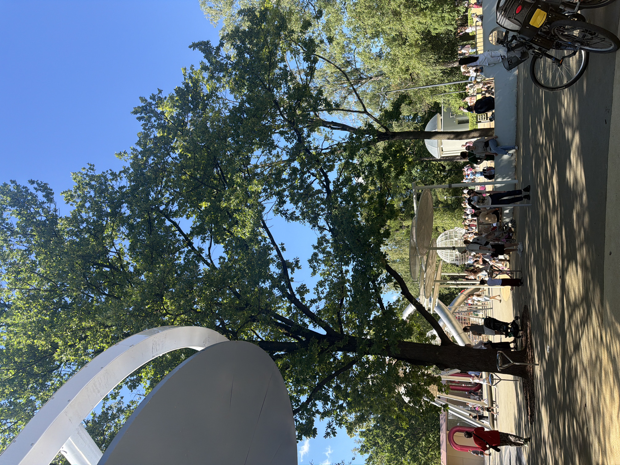 In the city of Unknown, Unknown, on a sunny day, a park scene unfolds. Large trees with lush green foliage dominate the frame, their branches casting dappled shadows on the paved ground. In the foreground, a modern, curved, white and gray architectural structure partially obscures the view of a playground.
The playground features silver tubular slides, geometric structures, and what appears to be a globe-shaped element. A diverse group of people, including adults and children, are present. Some are walking, others are standing or sitting on a low wall. Several individuals are gathered around a musical instrument, possibly a drum. Strollers and bicycles are also visible. The sky is a clear, vibrant blue.