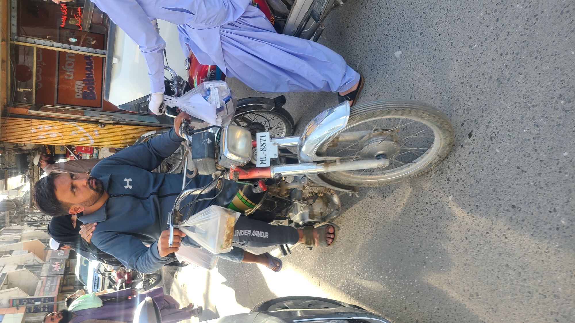 Here is a detailed description of the image:
**Scene Overview:** The image appears to be taken on a street in Quetta City Tehsil, Pakistan. The street is lined with shops and buildings. A man is sitting on a motorcycle, with a passenger behind him. There are other people and vehicles present in the vicinity.
**People:**
* **Man on Motorcycle:** He has a beard and is wearing a blue jacket and athletic pants. He is holding plastic bags containing items.
* **Passenger:** The passenger sits behind the man on the motorcycle.
* **Other People:** There are other people walking on the sidewalk and standing nearby.
**Objects:**
* **Motorcycle:** An old motorcycle with a license plate number "ML-8871."
* **Plastic Bags:** The man on the motorcycle is holding plastic bags containing items, possibly food.
* **Shops:** There are shops on the side of the street, including a shop with signs in Urdu.
* **Vehicles:** There are cars and other motorcycles on the road.
**Environment:**
* **Street:** The street is paved and appears to be busy.
* **Buildings:** There are buildings on both sides of the street, including shops and residential buildings.
**Actions:**
* The man is sitting on the motorcycle.
* He may be purchasing or delivering items.
**Interactions:**
* The man on the motorcycle is interacting with another person.