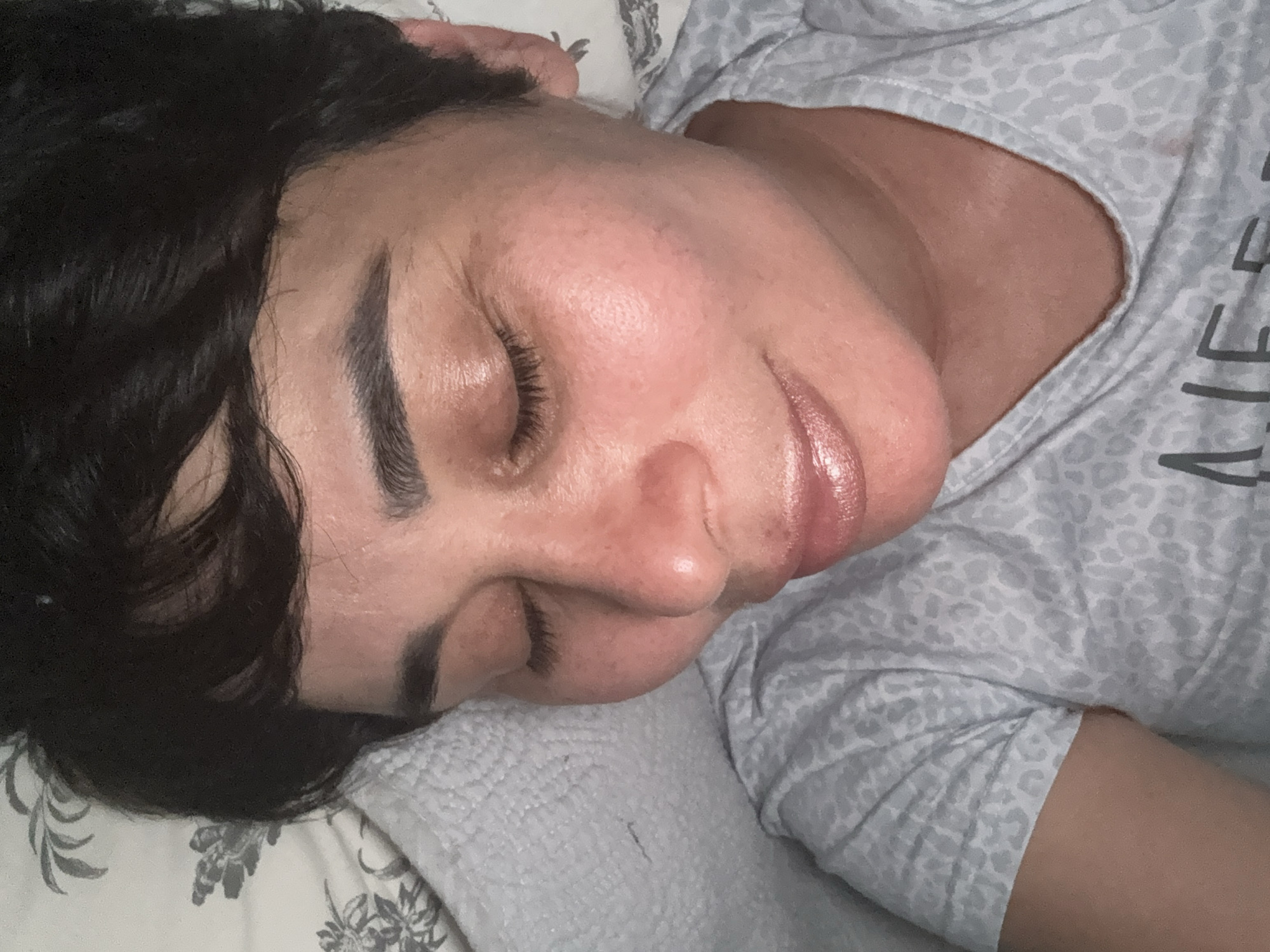 The image shows a close-up, tilted view of a person lying down with their eyes closed. The person has dark hair and is wearing a light-colored shirt with a leopard print pattern. The background consists of a light-colored patterned fabric, possibly a pillow or bedding. The lighting appears to be artificial and soft. The image does not contain any visual information that would allow for the determination of its location within the city of Bellmore, United States of America.