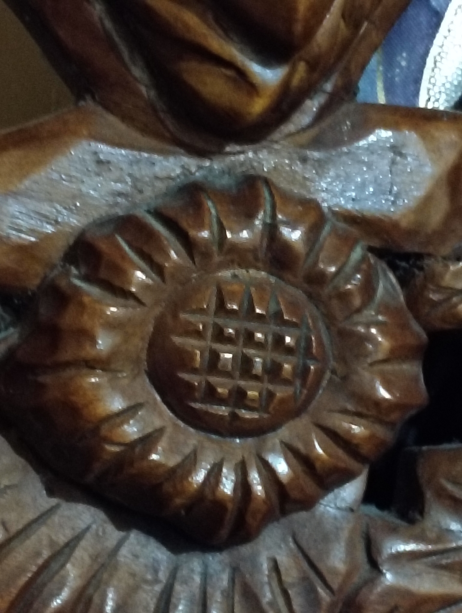 The image displays a close-up, detailed view of a carved wooden object, possibly part of a larger sculpture or furniture piece. The carving features a central circular motif with a grid-like pattern inside, surrounded by what appear to be stylized petals or leaves. The wood is dark brown and exhibits a glossy finish, highlighting the texture and depth of the carving. The background is out of focus, with hints of other carved wooden elements and possibly a textured surface. The lighting emphasizes the three-dimensional nature of the carving, creating shadows and highlights that define its intricate details. The scene is static and focuses solely on the craftsmanship of the wooden artifact.