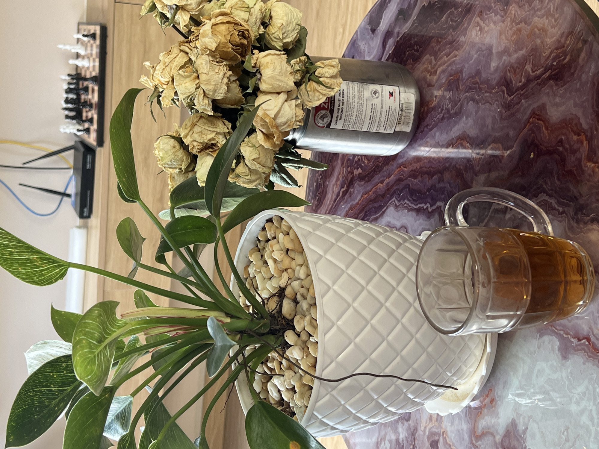 A high-angle shot captures a still life on a mottled purple and white surface, possibly a table. A white, diamond-patterned pot filled with light-colored pebbles and a green plant occupies the lower left quadrant. Next to it, a glass mug filled with a brown liquid, possibly beer or tea, lies on its side. A cylindrical metal container, acting as a vase, holds a bouquet of dried roses. In the background, partially visible on the left, are a chessboard and a router.