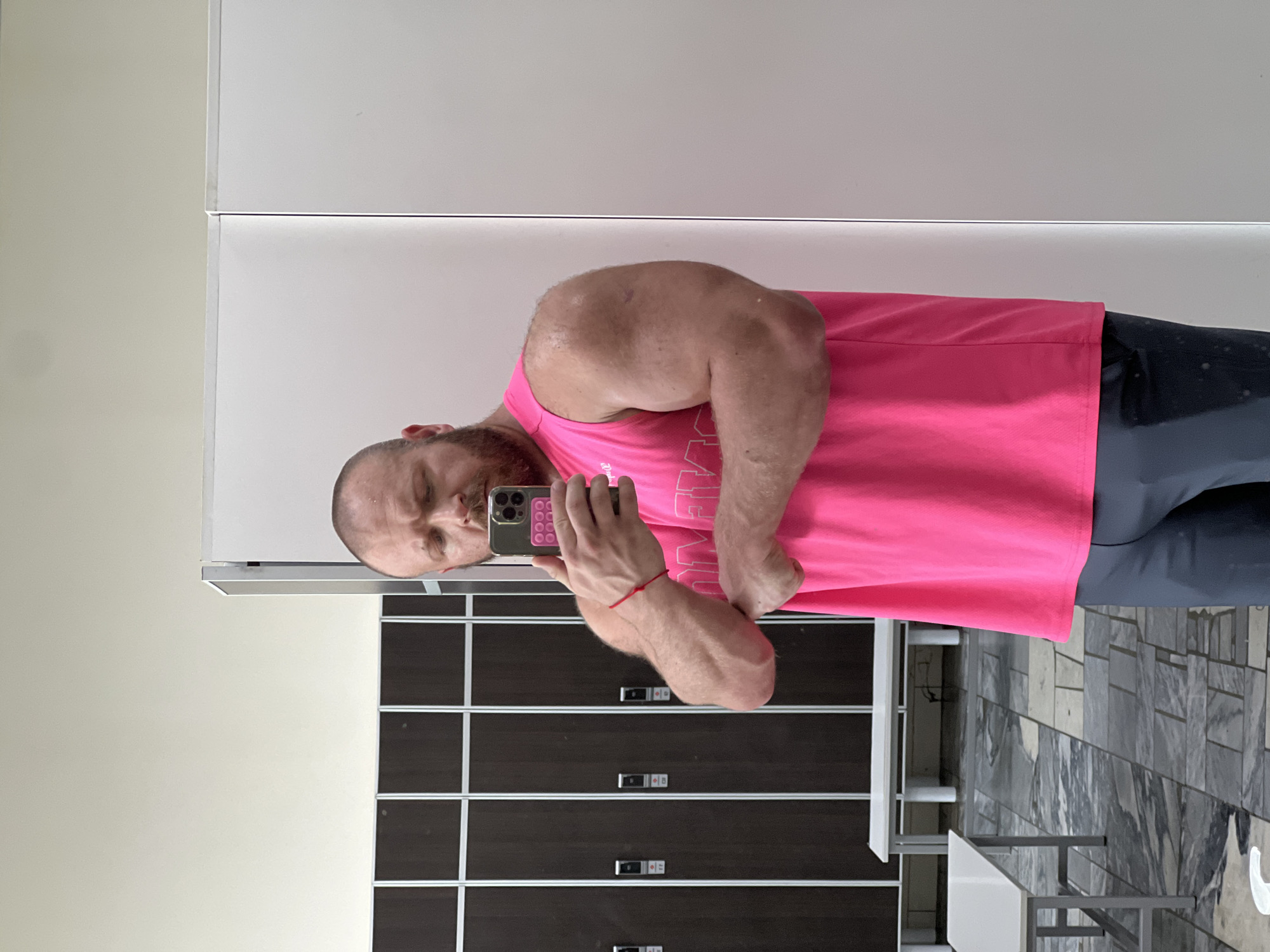 A fair-skinned male with a beard, wearing a bright pink tank top and grey pants, is taking a mirror selfie in what appears to be a locker room. He is flexing his bicep and looking at his reflection. The background consists of dark-colored lockers with silver trim, a white bench, and a tiled floor. The scene is brightly lit, suggesting an indoor environment. The location is identified as the city of Chaikovsky, Russia, based on metadata.