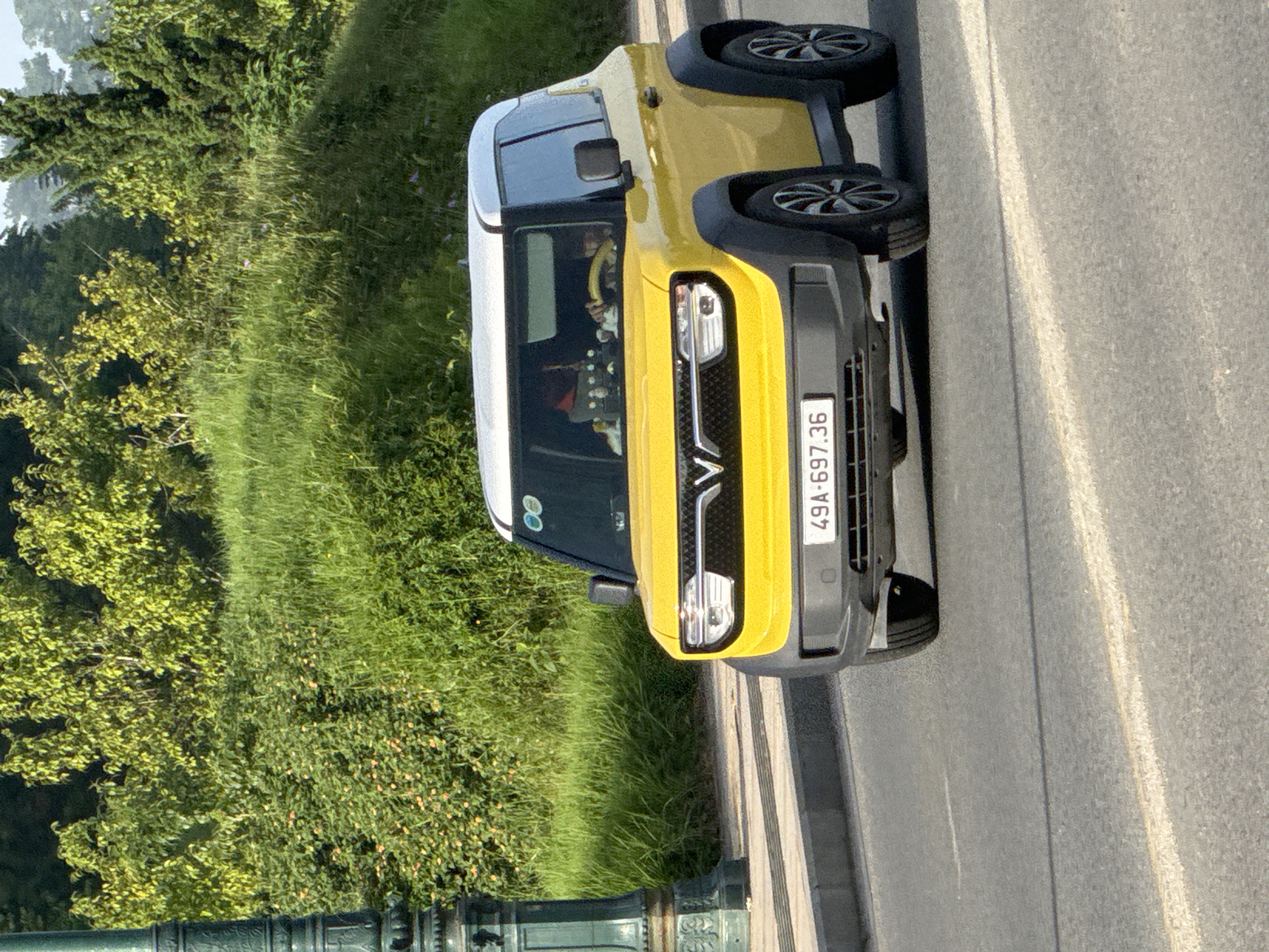 A yellow VinFast VF 3 electric car is driving on a paved road in Da Lat, Vietnam. The car has a white roof and black wheel wells. A person is visible driving the car. The car's license plate reads "49A-697.36". In the background, there is a grassy hill with trees and bushes. A dark green lamppost is visible on the left side of the image. The sky is overcast.