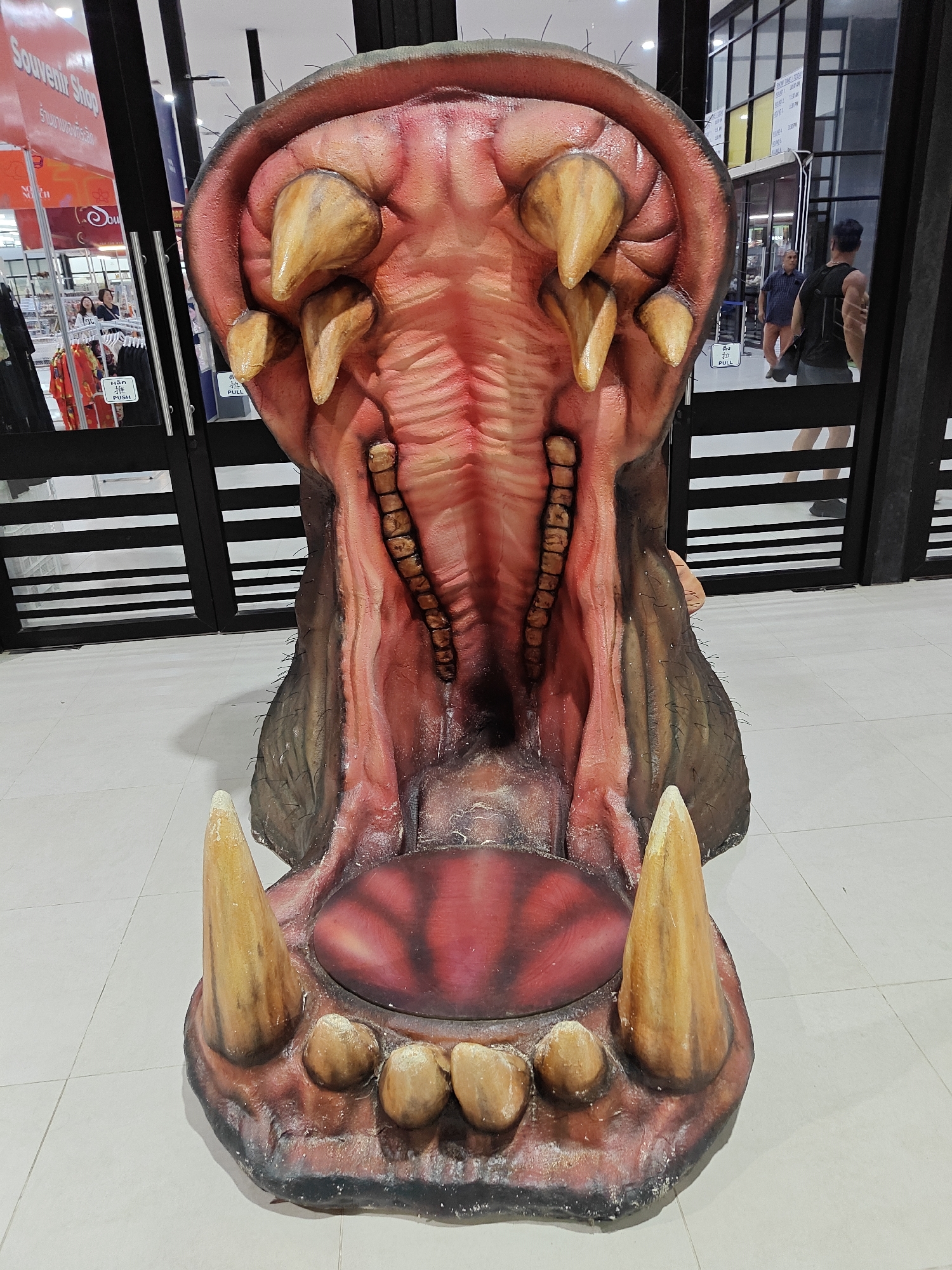 The image shows a large, realistic replica of an open hippopotamus mouth, likely a roadside attraction or photo opportunity, located outside a building with black and white striped gates. The mouth is detailed, with prominent teeth, a red tongue, and visible gums. Behind the hippo replica, a building with glass doors and windows is visible, with several people inside and outside. One person is wearing a black tank top and shorts, while another is wearing a blue checkered shirt and orange shorts. The ground is tiled, and a sign that reads "Souvenir Shop" is visible on the left. The overall scene suggests a commercial area, possibly a tourist spot, in Na Chom Thian, Thailand.