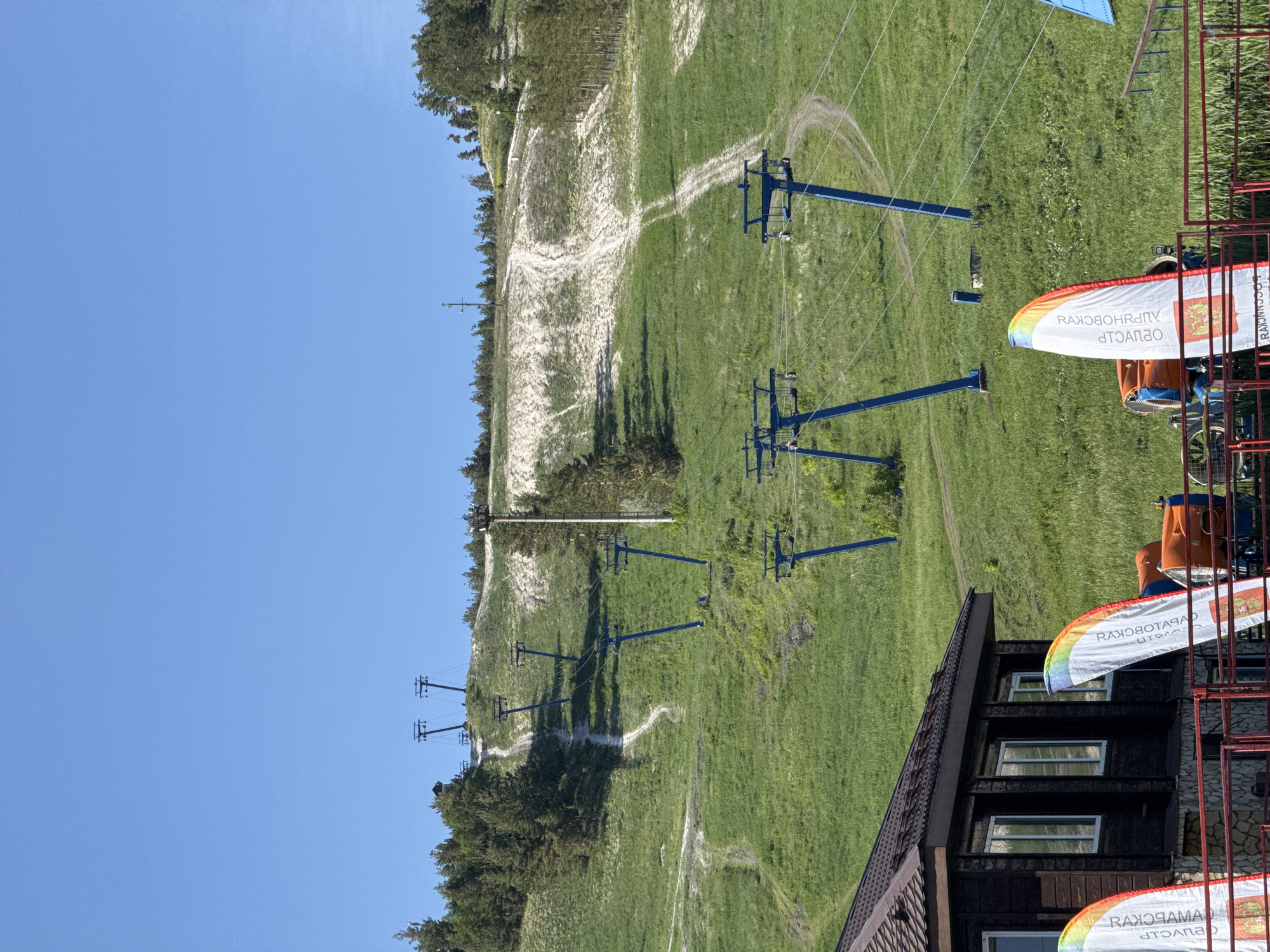 The image shows a ski resort in Khvalynsk, Russia on a sunny day. A series of blue ski lift support towers ascend a grassy hill towards a sandy area near the top. A dirt path winds alongside the towers. The base of the ski lift features a multi-story building with dark siding and a brown roof. Decorative banners with reversed Russian text are attached to the building's structure. The sky is clear blue.