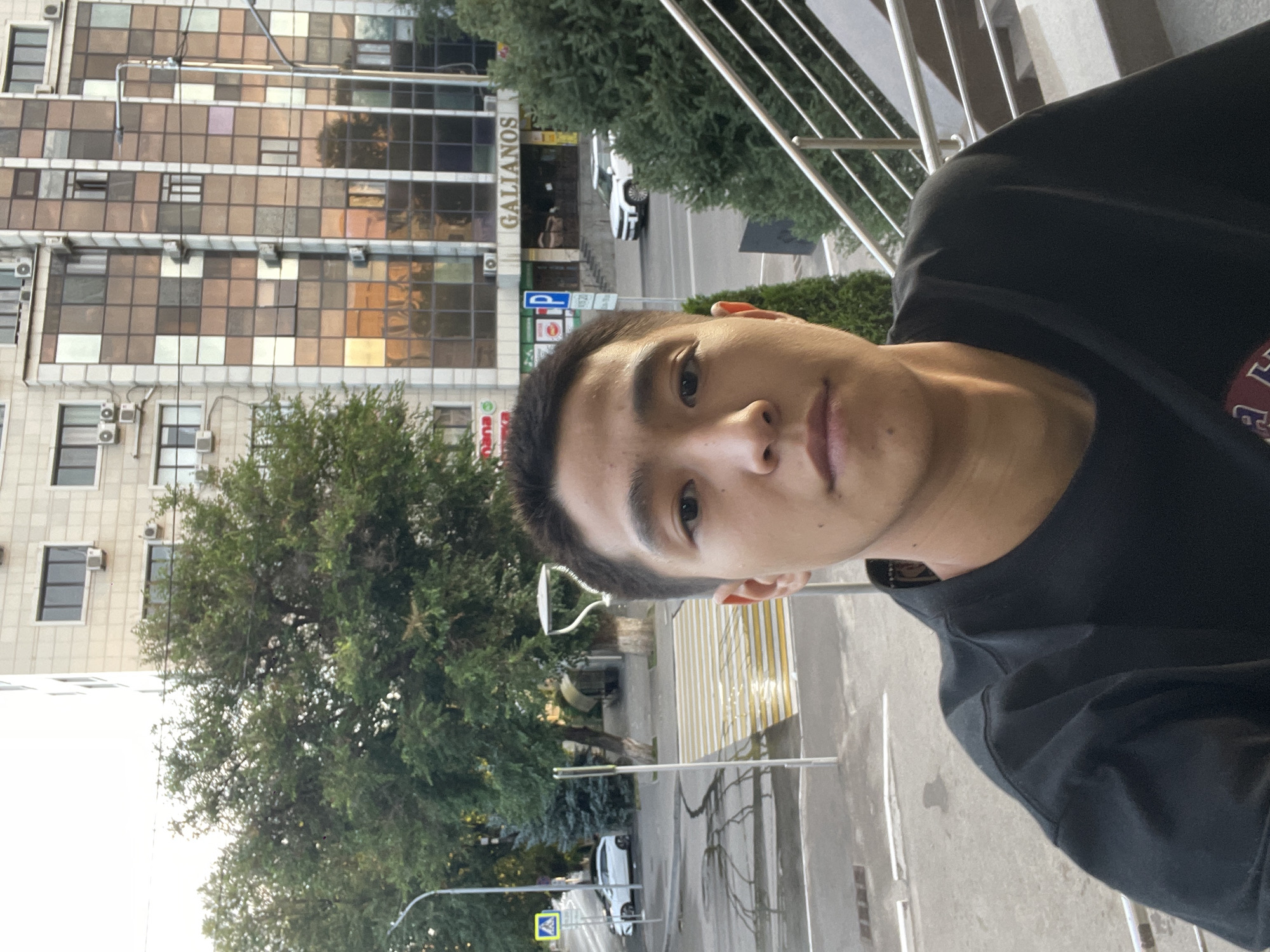 A medium shot photo captures a young Asian man in a black t-shirt, taking a selfie outdoors. He is positioned in the foreground, with a building and a street scene in the background. The building is a multi-story structure with a facade of glass windows in varying shades of brown and white, and air conditioning units visible. A sign on the building reads "GALIANOS." The street in front of the building features a pedestrian crossing with yellow and white stripes, a street light, and trees. A white car is visible on the street to the left. The overall environment suggests an urban setting in Almaty, Kazakhstan. The lighting indicates it is either early morning or late afternoon.