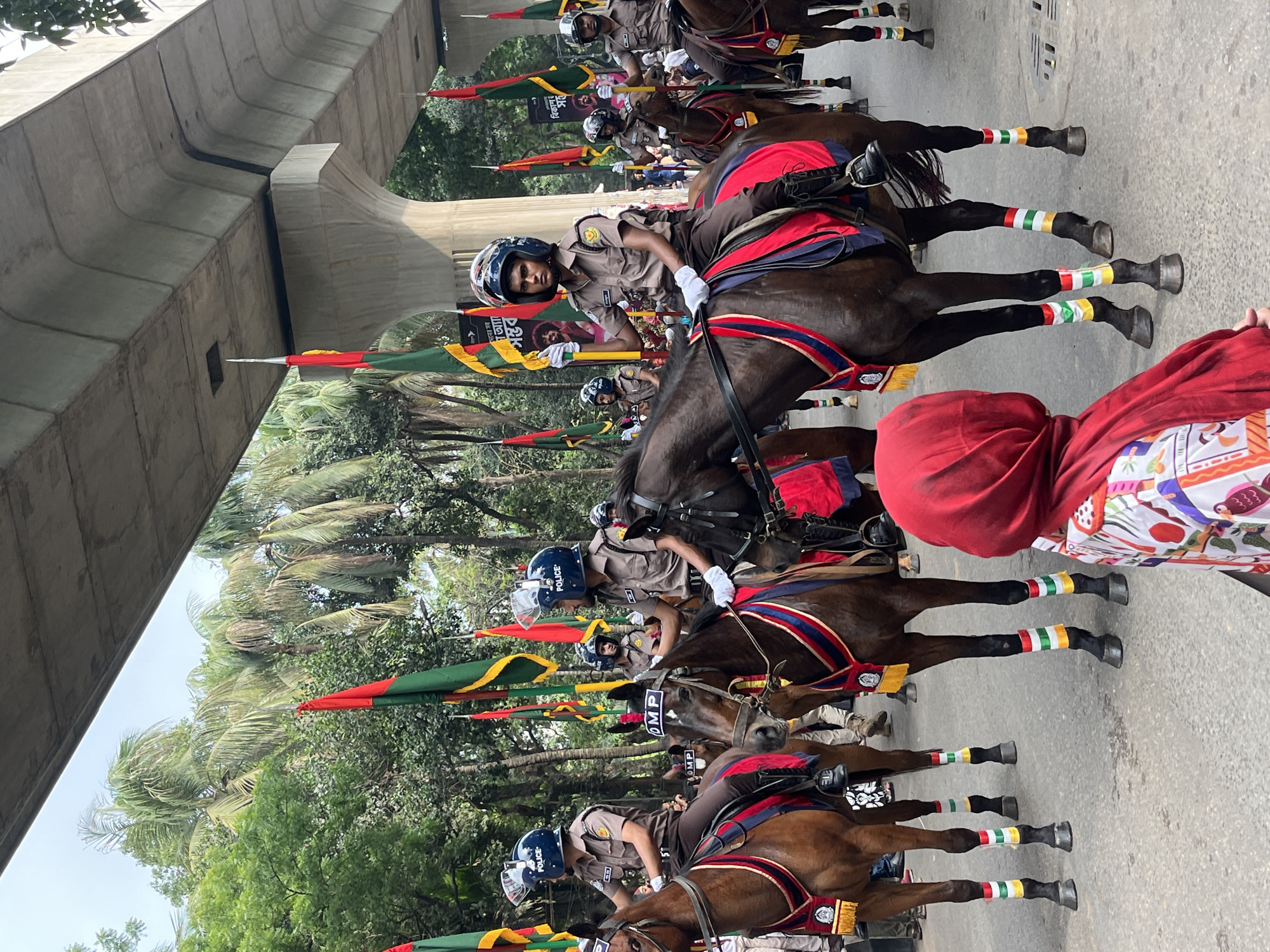 A mounted police parade is underway on a street in Dhaka, Bangladesh. The mounted officers, in khaki uniforms and blue helmets labeled "POLICE," ride dark horses decorated with red and blue trimmings and distinctive colored leg wraps of green, white, and red. They carry red and green flags.
The parade is positioned under a concrete overpass. Trees and vegetation are visible in the background. In the foreground, a person wearing a red hooded garment with colorful patterns looks on.