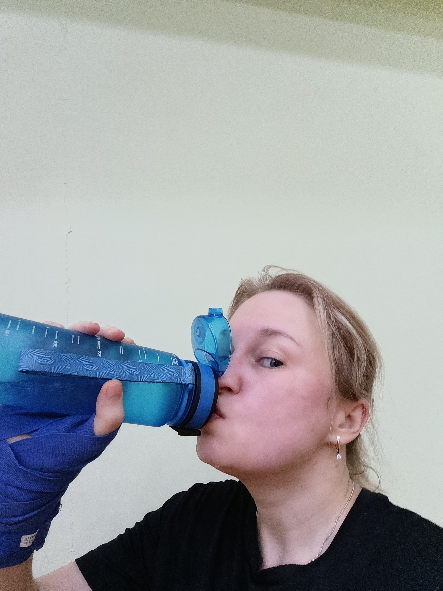 A medium shot captures a fair-skinned woman drinking from a blue water bottle. She wears a black t-shirt and has blond hair. Her left hand is wrapped in a blue bandage. A silver necklace adorns her neck, and she sports a drop earring. The background is a plain, off-white wall. The location is not specified.
