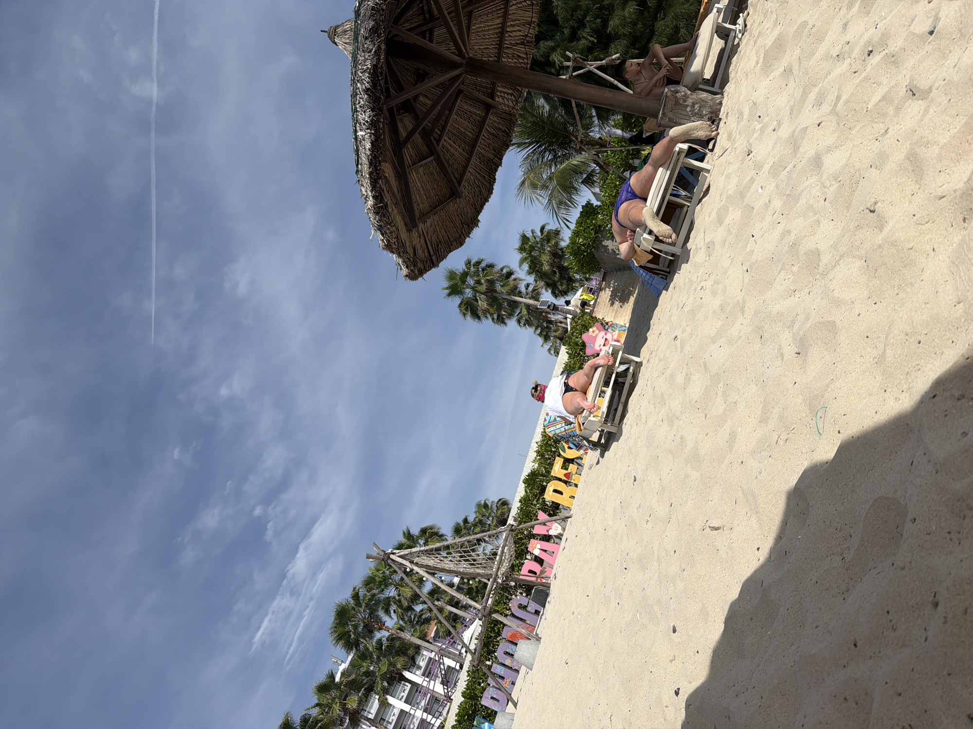 The image depicts a sunny day at a beach, possibly in Dong Ninh Hoa Ward, Vietnam, as suggested by the "PHUOC LONG BAY RESORT" sign. In the foreground, the sandy beach slopes upwards with a large shadow cast by an unseen object. Several people are relaxing on lounge chairs. One person, wearing a white top and black bikini bottoms, is lying on a chair to the left of the sign. To the right, under a thatched umbrella, another person in a blue bikini is lying on a lounge chair, with their back to the camera. Further to the right, a young man in black shorts is sitting on a lounge chair. In the background, palm trees and a multi-story building with purple accents are visible. The sky is blue with scattered white clouds and a faint contrail from an airplane. The overall atmosphere is one of leisure and relaxation at a tropical resort.