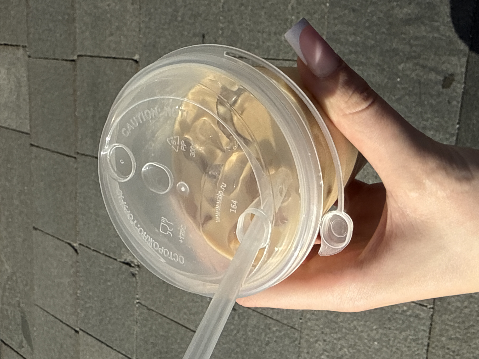 A high-angle medium shot shows a person holding a clear plastic cup filled with iced coffee. The cup has a transparent lid with a straw inserted. Text is visible on the lid, including "CAUTION" in English and "ОСТОРОЖНО. ГОРЯЧ" in Russian. The lid also features a symbol of a cup with steam and the text "+120C". Below that, the recycling symbol for "PP" and the number "300" are present. Further text on the lid includes "www.vzljp.ru" and the number "164". The background is a grey, textured pavement with visible grout lines. The person's hand and fingernails, with a French manicure, are also in the frame. The lighting suggests it is daytime, casting a shadow from the cup onto the pavement. The presence of Russian text on the cup indicates the location is likely in Moscow, Russia.
