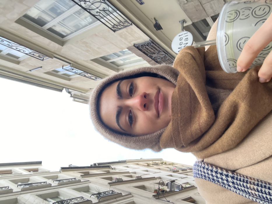 The image displays a low-angle selfie of a young woman centered in a narrow street in Paris, France. The subject has dark hair, dark eyebrows, brown eyes, and fair skin, with a subtle smile. She wears a light brown hooded garment, a thick camel-colored scarf wrapped around her neck, and a beige overcoat. A blue and white houndstooth patterned bag strap is visible over her left shoulder. Her right hand holds a clear plastic cup with a lid and straw, adorned with a pattern of black outline smiley faces. The cup contains a light green beverage. The background consists of tall, light-colored stone buildings consistent with Haussmannian architecture, featuring multiple windows and decorative wrought-iron balconies on both sides of the street. The sky above is bright white and overcast. On the building to the subject's right, a circular white sign with "Meet Nail" in black text is visible, alongside a partially obscured blue street sign. A traditional multi-branched street lamp is affixed to the building on the left.