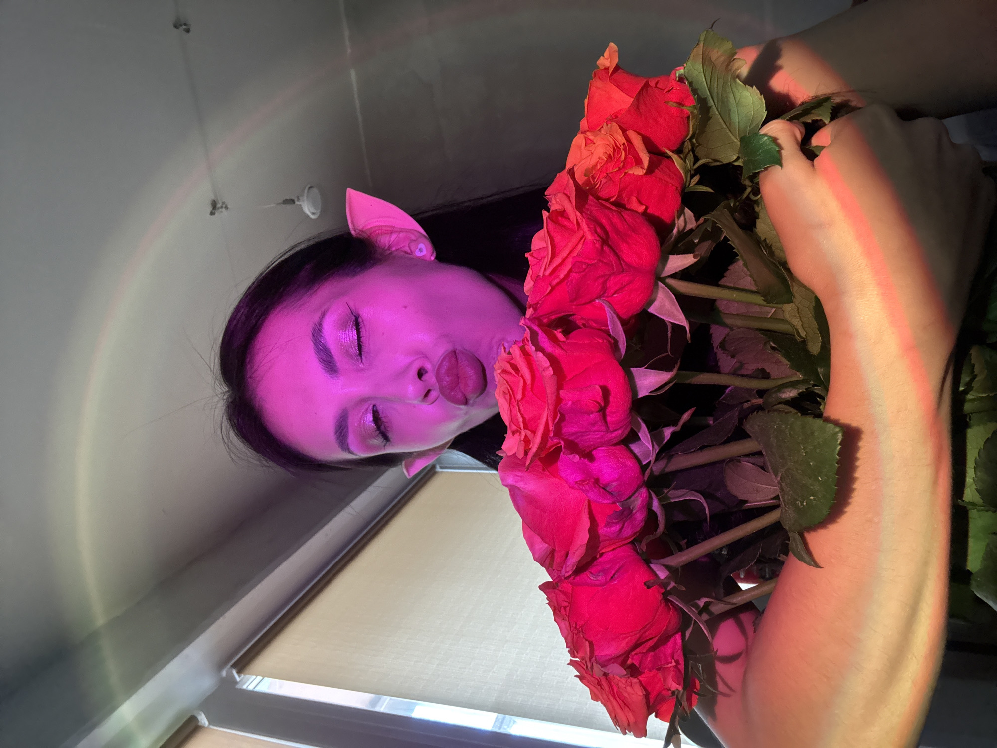 The image, rotated 90 degrees counter-clockwise, depicts a stylized portrait of a person holding a large bouquet of roses indoors in Votkinsk, Russia. The subject is a young person, likely female, with dark hair, visible from the shoulders up. Their face and parts of their arms are illuminated by a vibrant magenta and pink light, creating a surreal and artistic atmosphere.
The person is captured looking upwards with their eyes closed and their tongue playfully extended. They wear subtle, pointed light-pink elf ears, adding a whimsical touch to their appearance. Visible makeup, including sparkling eyeshadow, enhances their features under the colored light.
They are holding a substantial bouquet of red and orange roses, with green leaves and stems clearly visible. The same pinkish-red light that illuminates their face also casts streaks onto their arm as they hold the flowers.
In the background, a light-colored wall or ceiling features a subtle, multi-colored rainbow-like light refraction pattern, suggesting sunlight entering the room and interacting with a prism or similar object. Part of a window or a window blind is visible towards the lower left (when the image is rotated to a standard upright view), confirming an indoor setting. The photograph is taken from a low angle, looking up at the subject. The overall impression is one of a playful, dreamlike, or fantasy-themed photoshoot. There is no visible text in the image.