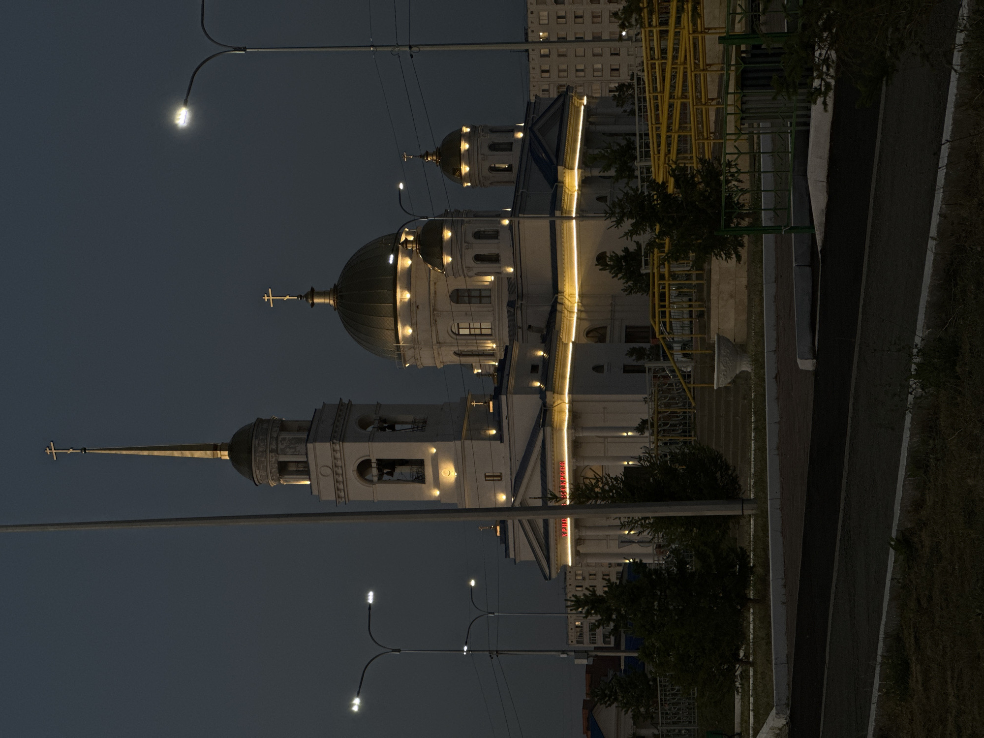 The image depicts the Holy Ascension Cathedral in Kokshetau, Kazakhstan, at dusk or night. The white cathedral features a tall bell tower topped with a golden spire and cross, and several golden domes, also adorned with crosses. Yellow lights illuminate the architectural details of the building, including the edges of the roofline and around the windows and entrance. A large red inscription is visible above the main entrance. Surrounding the cathedral are several evergreen trees, streetlights with bright white bulbs, and a yellow metal railing system, suggesting the presence of a pathway or ramp. In the background, apartment buildings are visible, illuminated by some interior lights, and power lines cross the sky. The foreground shows a dark asphalt surface with a concrete curb and grassy areas. There are no people visible in the image.
