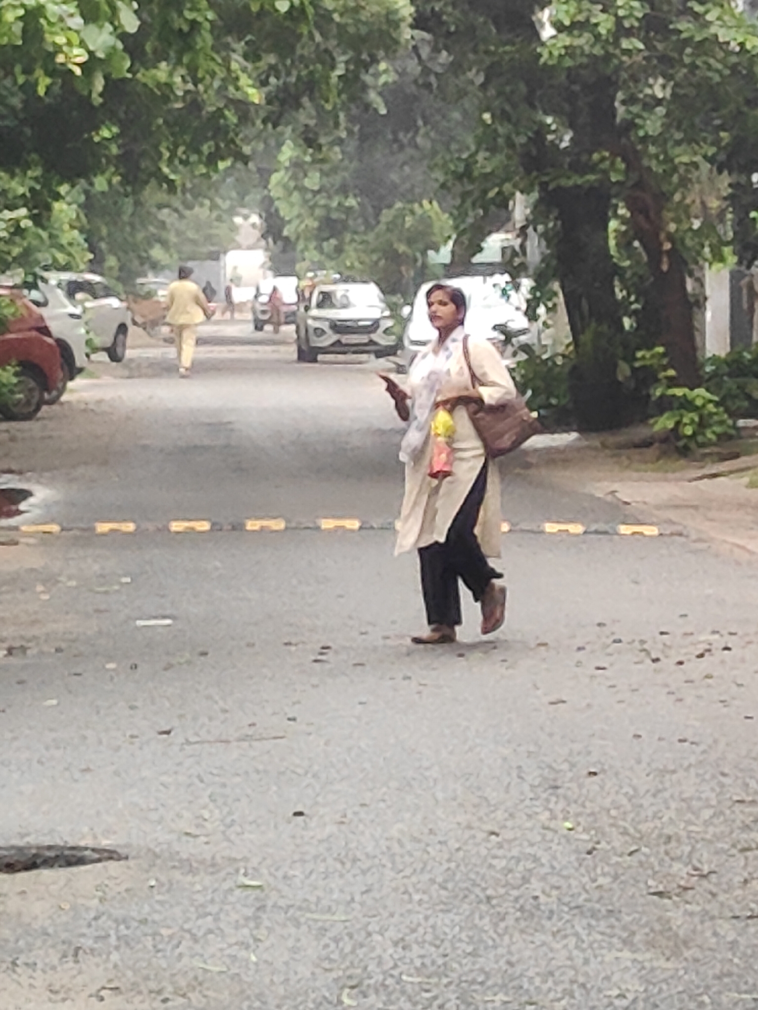 A woman in a cream-colored tunic and dark pants walks across a wet asphalt street in Gurgaon, India. She carries a brown bag over her right shoulder and holds a smartphone in her left hand. Yellow speed bumps are visible across the road. Trees line the street, and parked cars are present on the left side of the image. The sky appears overcast, and the overall scene suggests it may have recently rained.