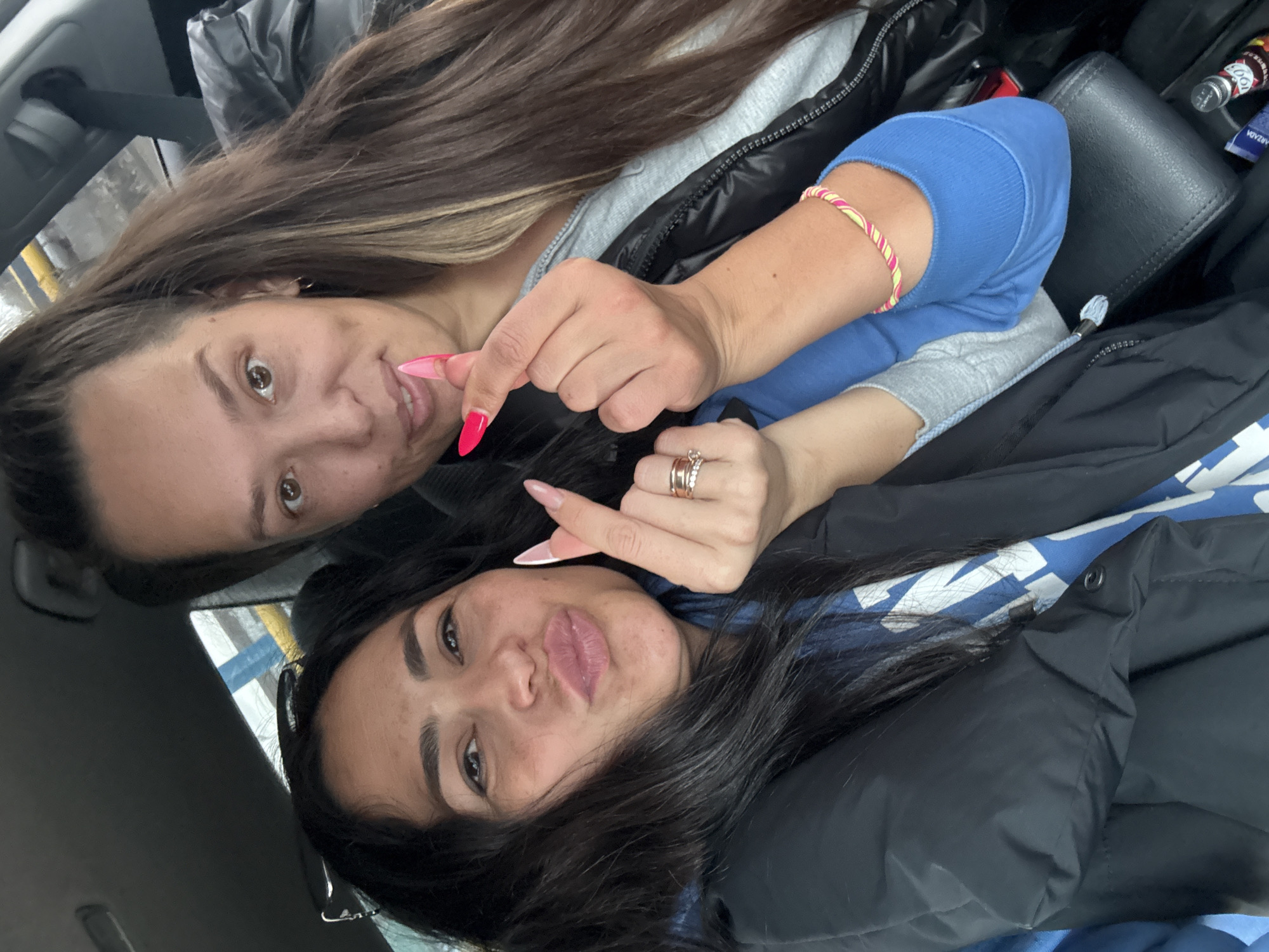 Two women are captured in a close-up, diagonal portrait, likely a selfie, inside what appears to be a car in Chaikovsky, Russia. Both are looking towards the camera with casual expressions.
The woman in the foreground has long dark hair and is wearing a black puffer jacket over a blue top with some indistinct white text. She has long, light pink or white nails and is making a half-heart gesture with her thumb and index finger, showing multiple rings on her ring finger. Her lips are slightly pursed.
Behind her, partially obscured, is another woman with long brown hair, wearing a black puffer jacket over a grey hooded garment and a blue long-sleeved top. She has bright pink/red long nails and is also making a half-heart gesture with her fingers, mirroring the gesture of the other woman. A striped pink and yellow bracelet is visible on her left wrist.
The background shows elements of a car interior, including a seatbelt strap and glimpses of a dashboard or door panel. On the car seat next to the women, a black purse or wallet is visible, along with a small silver bottle partially labeled "100" and a tube with "ARZADA" written on it. The overall mood is casual and friendly, with the heart gestures suggesting affection or solidarity.