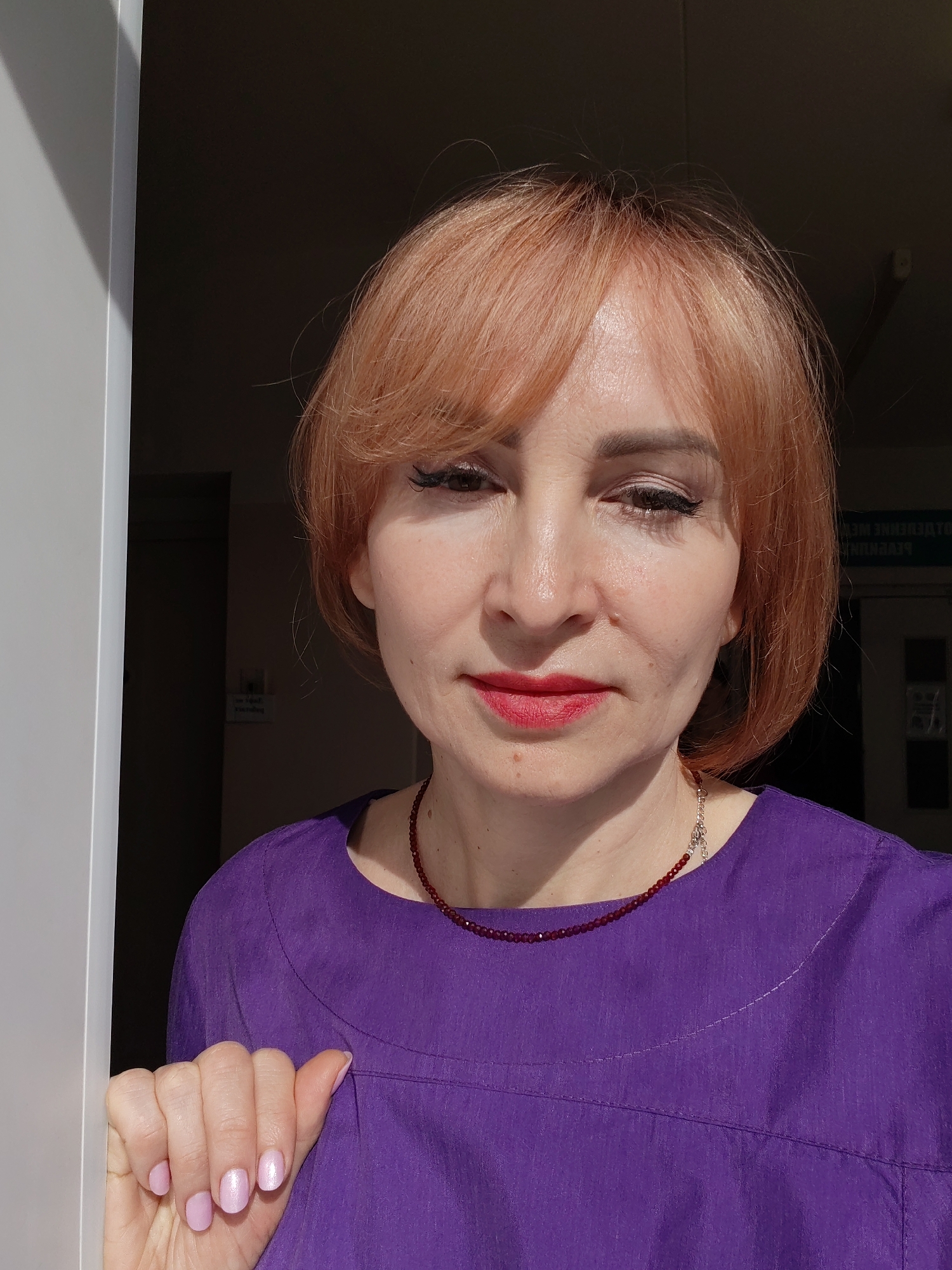 A woman with short, reddish-blonde hair and fair skin is seen in a close-up shot, partially obscured by a white door frame on the left. She is wearing a purple top and a red beaded necklace. Her makeup includes eyeliner and red lipstick. The background is dark, with indistinct shapes suggesting an indoor space, possibly a doorway leading inside. A partially visible sign or label with reversed Cyrillic text can be seen on the right side of the image, indicating the location might be in Russia. Based on this and given the prompt to include the location of Chaikovsky, Russia, it is likely the location is within that city.