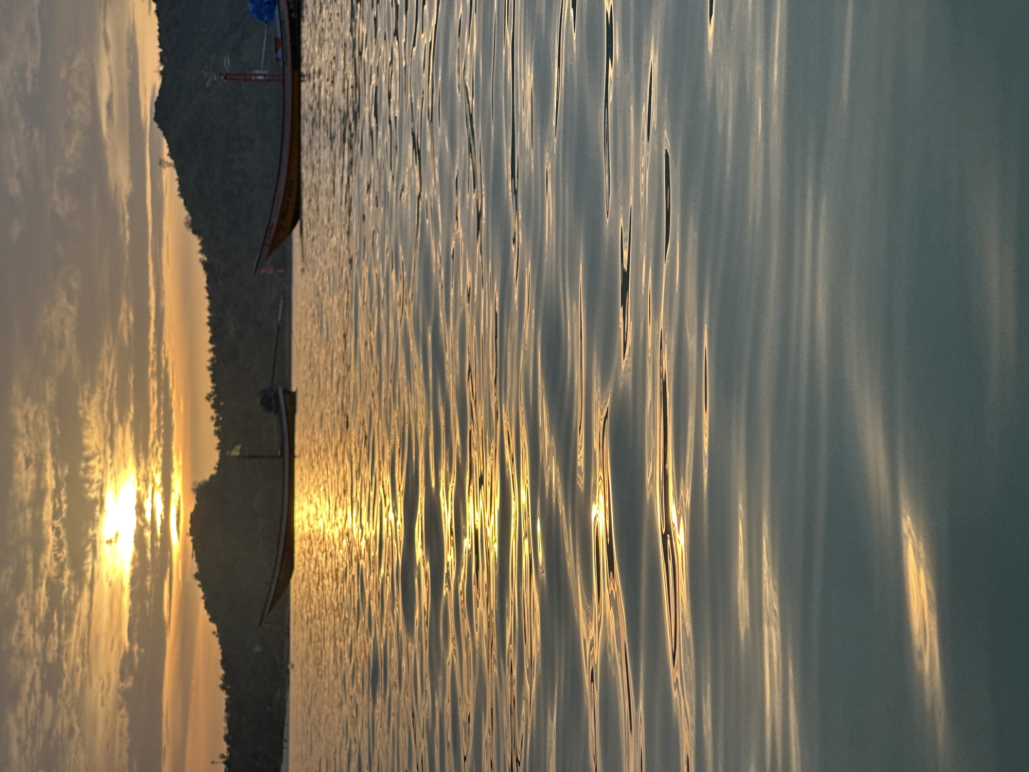 The image depicts a serene seascape at sunset in Ko Pha-ngan District, Thailand. The sun, a brilliant orb of orange, is partially obscured by clouds, casting a warm, golden glow across the sky and reflecting on the rippling water. The water's surface is a mosaic of gentle waves, catching the sunlight and creating streaks of light and shadow.
In the middle ground, two longtail boats are anchored in the calm sea. The boat on the left is closer to the shore and appears to be empty. The boat on the right is further out, also appearing unoccupied. Both boats are typical of the region, with their distinctive slender hulls and overhead structures.
Behind the boats, a silhouetted island rises from the sea. Its dark outline is softened by the twilight, and the lush vegetation that covers its slopes is visible as a dense texture against the sky. The island is framed by the warm hues of the sunset sky, with scattered clouds adding depth and drama to the scene.
The overall impression is one of tranquility and natural beauty, capturing a moment of peaceful repose as day transitions into night in this picturesque Thai district.