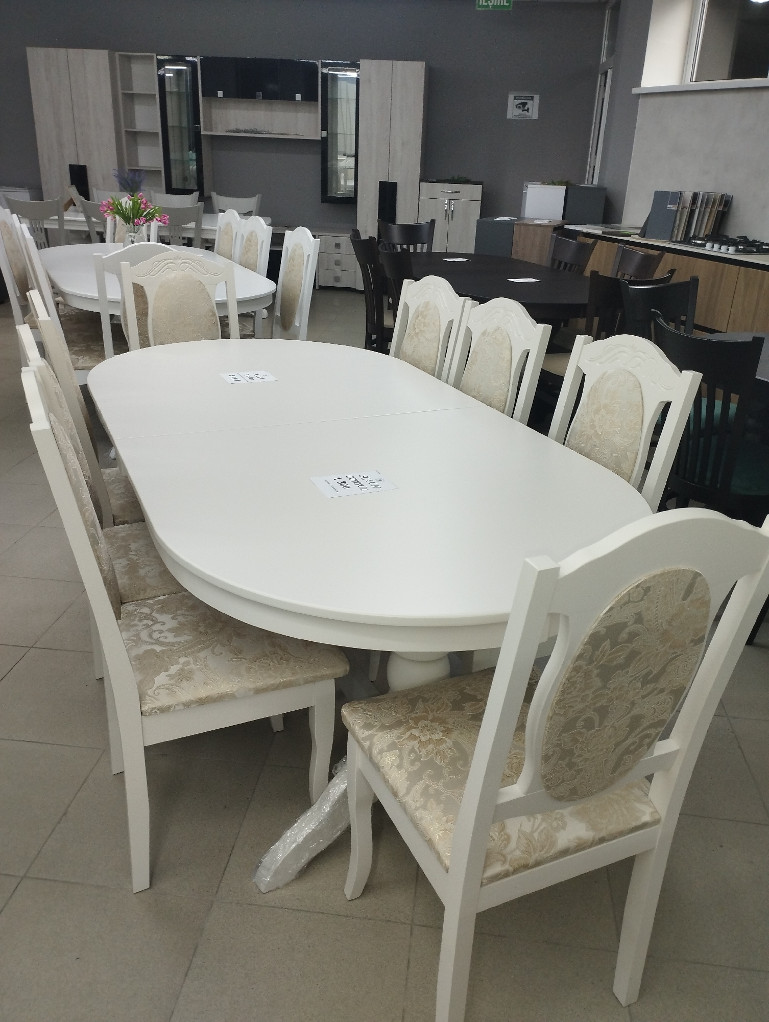 A white oval dining table is surrounded by six white chairs with ornate floral-patterned upholstered seats and backs. The table has a price tag attached. In the background, there are several other tables and chairs, including a dark wood dining set with chairs. The room appears to be a furniture showroom. The floor is tiled with light gray tiles. The walls are painted a dark gray color. There are also some cabinets and shelves visible in the background. This furniture showroom is located in the city of Fălești, Moldova.