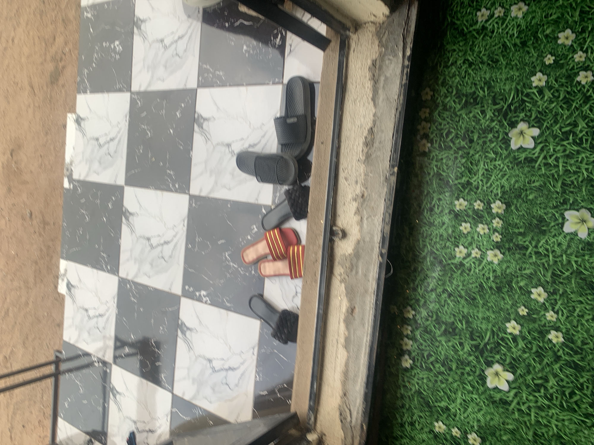 In Jauro Yinu, Nigeria, a tiled entryway features a collection of footwear. The floor is a checkered pattern of grey and white marble-effect tiles. To the left, a pair of black slides with a braided strap rests on the tiles. Next to them, a pair of red slides with gold stripes is positioned. Further to the right, two black slides, one with a textured sole, are placed. The entryway opens onto a bright green mat adorned with a pattern of white flowers. The scene suggests a domestic or commercial entrance.