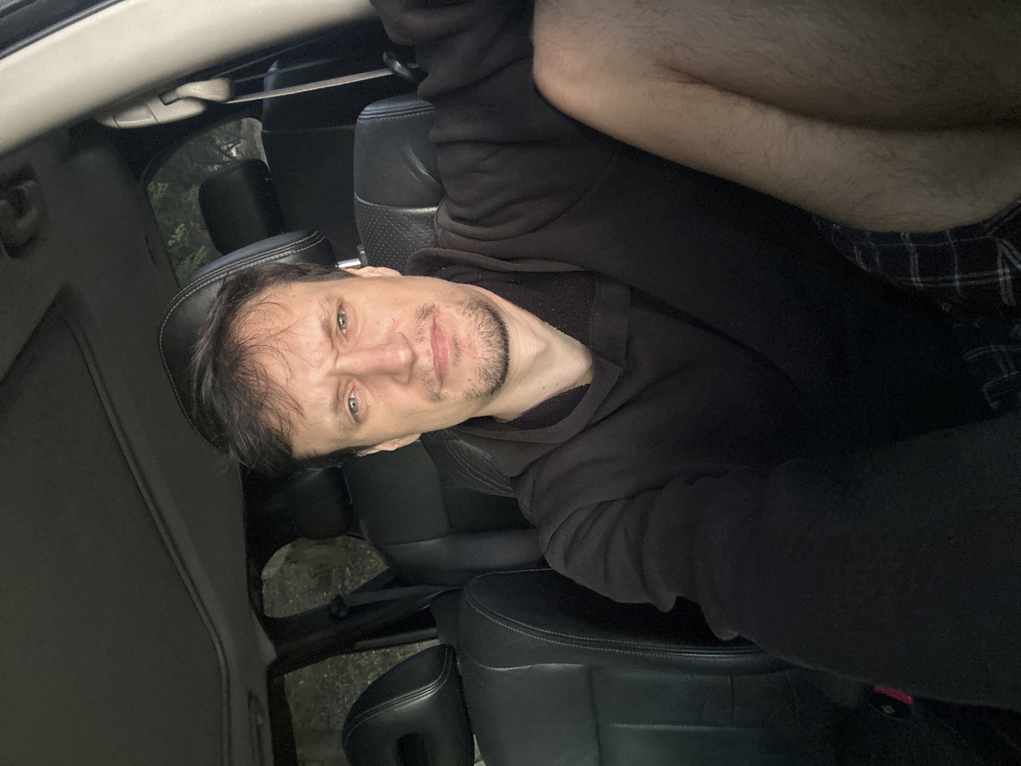 A man with a frowning expression sits in the driver's seat of a car, looking directly at the camera. He is wearing a black hooded sweatshirt and plaid shorts. The interior of the car is dark, with black leather seats visible. Outside the car, trees and foliage can be seen through the windows, suggesting an outdoor, possibly rural, setting. The location is identified as Makiv, Ukraine.