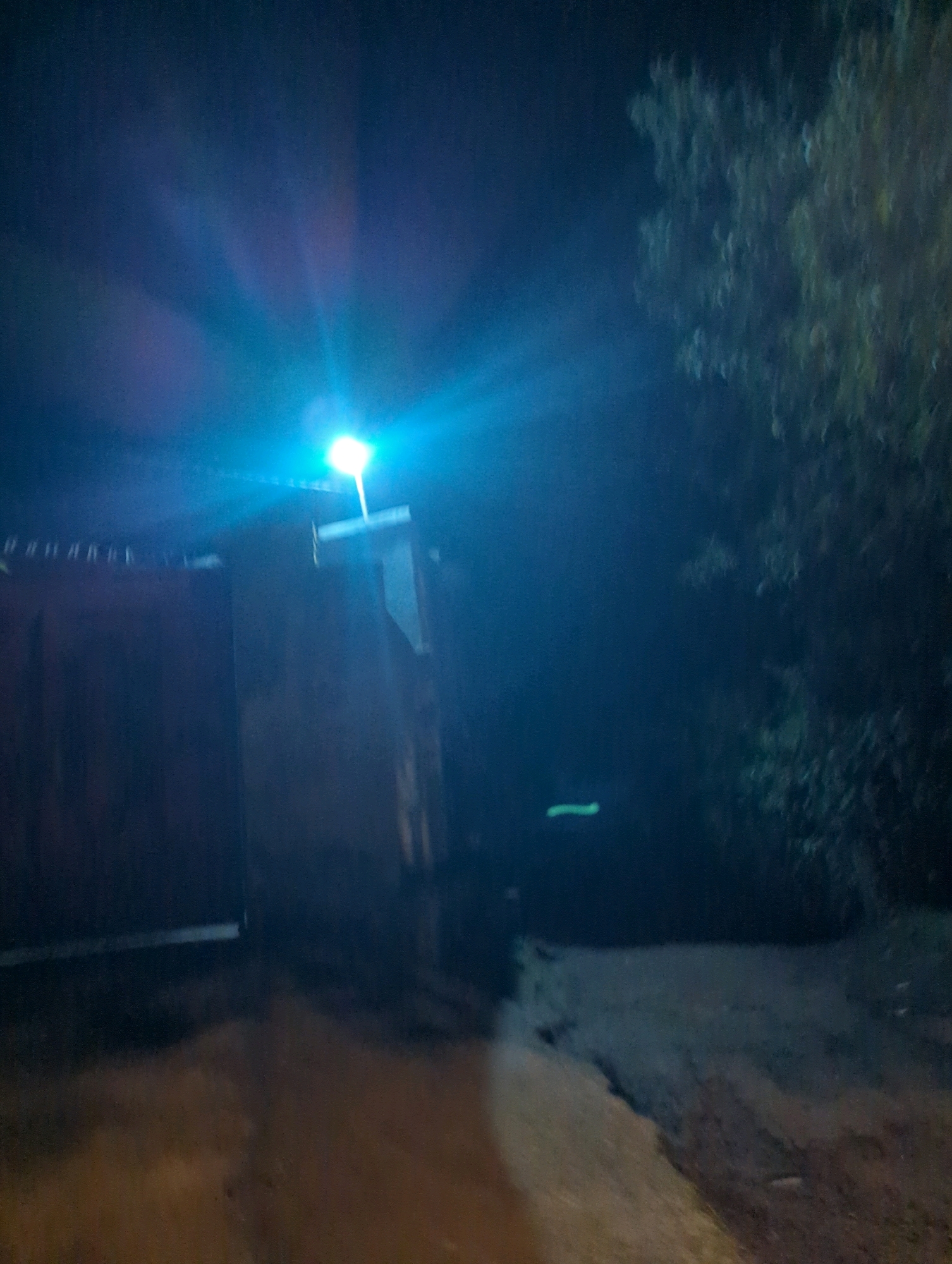 A night-time outdoor scene is depicted, illuminated by a bright blue-white street lamp situated above a dark, rectangular structure, possibly a gate or a wall. The light casts strong rays and creates a noticeable glare. To the right of the lamp, the silhouette of a tree with dense foliage is visible against the dark sky. The ground in the foreground appears to be a dirt path or road, unevenly lit by the street lamp and showing patches of light and shadow. The overall atmosphere is dark and somewhat hazy. The location is identified as Musanze District, Rwanda.