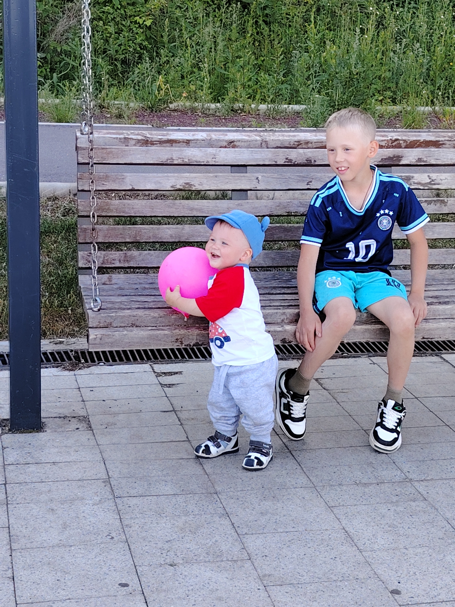 In the city of Chaikovsky, Russia, two boys are observed outdoors on a paved area adjacent to a wooden slatted bench. A younger boy, appearing to be a toddler, stands in the foreground on the left. He wears a blue cap, a white and red long-sleeved shirt with a graphic resembling a vehicle, light grey sweatpants, and black and white sandals. He holds a spherical pink object, likely a ball or balloon, with both hands and displays a wide smile, looking towards the left of the frame. An older boy, appearing to be a child between six and eight years old, is seated on the right side of the wooden bench. He wears a dark blue soccer jersey with light blue accents and the number "10", light blue shorts, olive-colored socks, and black and white athletic shoes. He is also smiling, with his gaze directed slightly to the left. Behind the bench, dense green foliage is visible, with a narrow paved strip bordering it. A vertical dark grey metal pole is positioned on the far left, and a metal chain, possibly part of a swing, hangs near the pole and over the left end of the bench. The ground consists of light grey square paver tiles, and a metal drainage grate is present directly beneath the wooden bench.