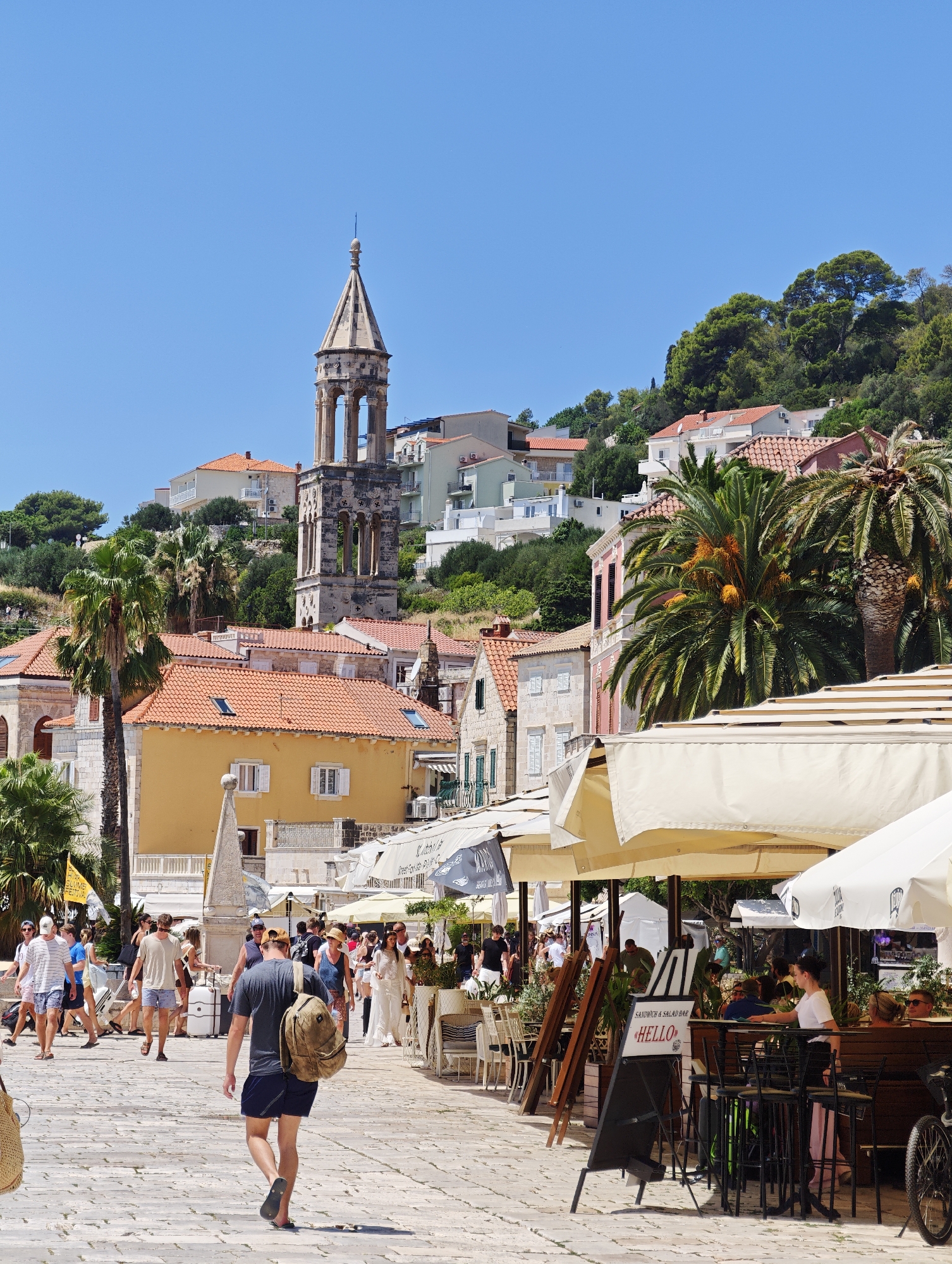 The image shows a sunny day in Hvar town, Croatia, specifically on a cobblestone promenade lined with shops and cafes. In the background, the historic bell tower of St. Stephen's Cathedral stands tall, overlooking a cluster of buildings with terracotta roofs. Palm trees are scattered throughout the scene, adding to the Mediterranean ambiance.
The promenade is bustling with activity. Tourists, dressed in casual summer attire, stroll along, some carrying shopping bags. Outdoor cafes with white umbrellas offer seating for patrons enjoying the pleasant weather. A man in a grey t-shirt and dark shorts, carrying a backpack, walks away from the camera. Another man in a blue t-shirt and shorts is seen walking towards the left. A woman in a white dress, possibly a bride, is seen in the background, walking with a man.
The shops and cafes feature various signs, including "SANDWICH & SALAD BAR HELLO." The overall scene depicts a lively tourist destination during the daytime.
