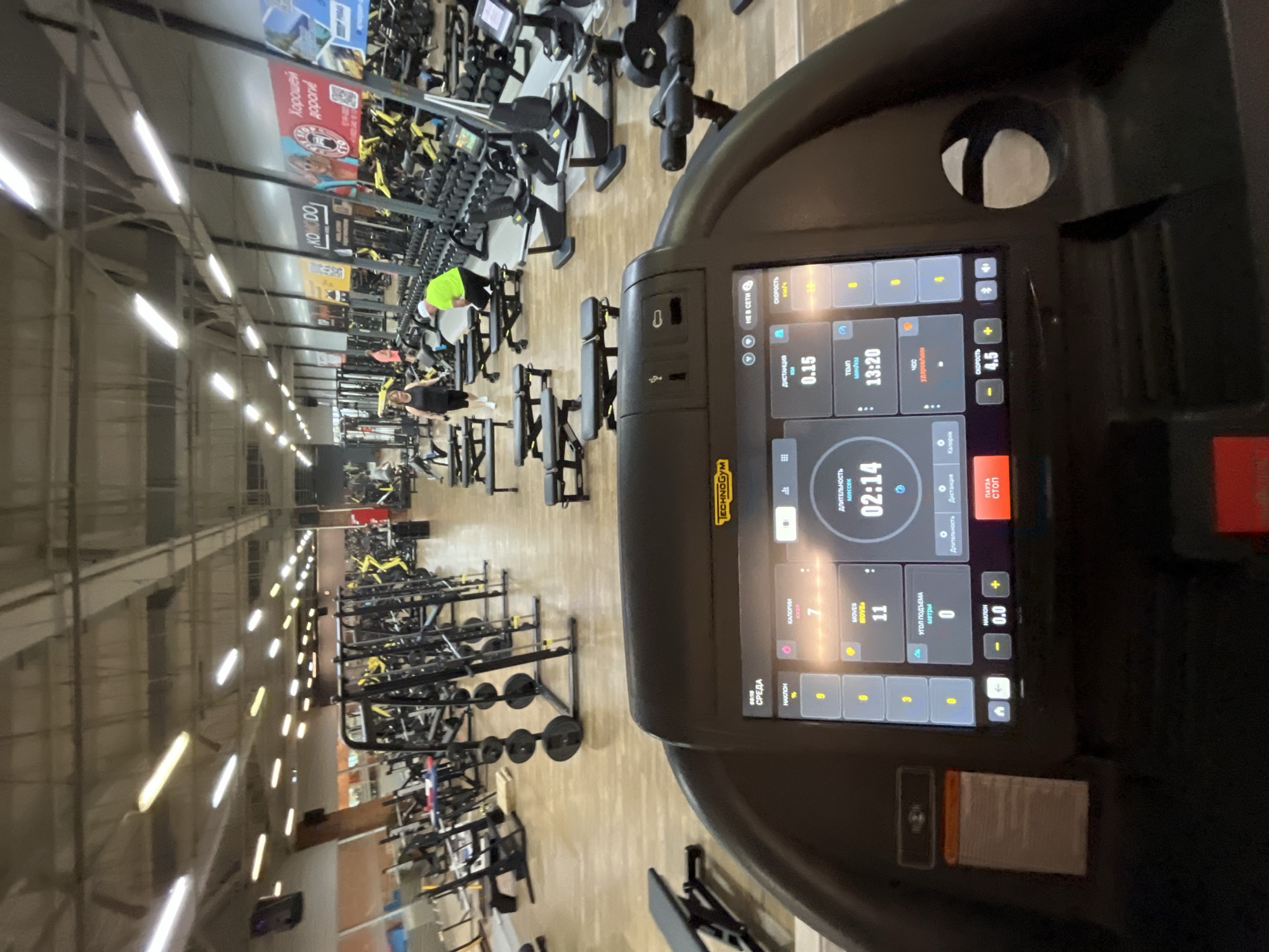 The image shows a treadmill in a gym. The treadmill's display shows the current time as 02:14, a distance of 0.15 km, a speed of 4.5 km/h, and the incline is set at 0.0. The screen also shows that 7 calories have been burned and 11 moves have been recorded.
The background of the gym includes weightlifting equipment, exercise benches, and several people working out. A sign saying "KOMODO, Хорошей дороги!" can be seen in the background. This text indicates the location to be in the city of Chaikovsky, Russia. The gym is well-lit with rows of fluorescent lights.
