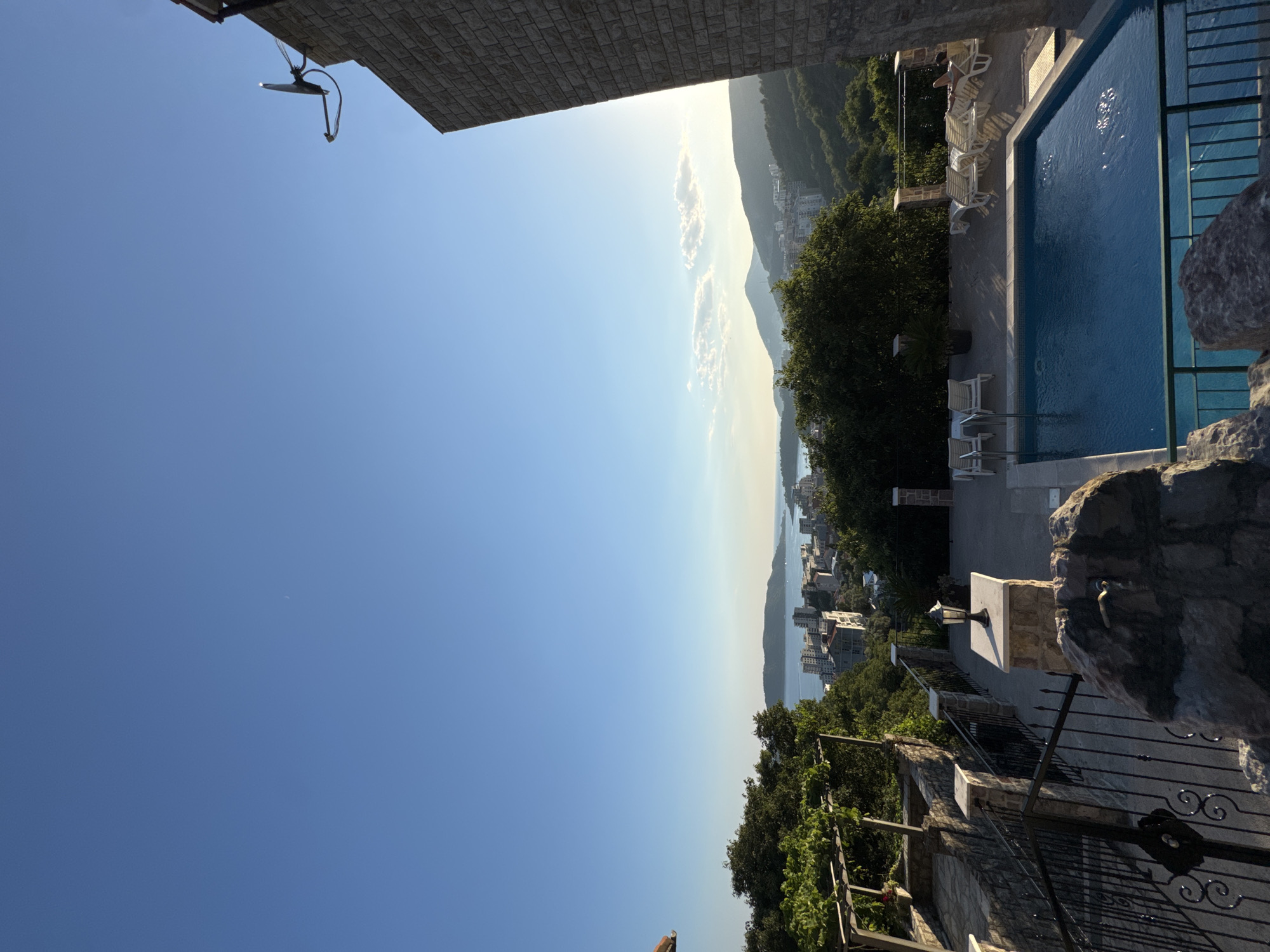 This is a photograph of a swimming pool area overlooking a coastal city and the sea, likely in Rafailovići, Montenegro. The sun is setting or rising, casting a warm glow on the scene.
In the foreground, a stone-clad building rises on the right side of the frame. A satellite dish is mounted on the exterior wall. Below, a swimming pool with clear blue water is in the center of the image. Several white lounge chairs are arranged around the pool deck, with some occupied by sunbathers. One person is visible reclining on a chair.
To the left of the pool, a terraced area with stone steps and railings leads down. There are decorative lamps and stone planters. Lush green trees and vegetation surround the property, with grapevines visible on a trellis.
In the background, the cityscape of Rafailovići unfolds, with buildings of varying heights clustered along the coast. Beyond the city, the calm sea stretches towards a distant, hazy horizon, with rolling hills and mountains forming the backdrop. The sky is a clear, bright blue, with a few wispy clouds near the horizon.
The overall impression is one of a serene and picturesque vacation setting.