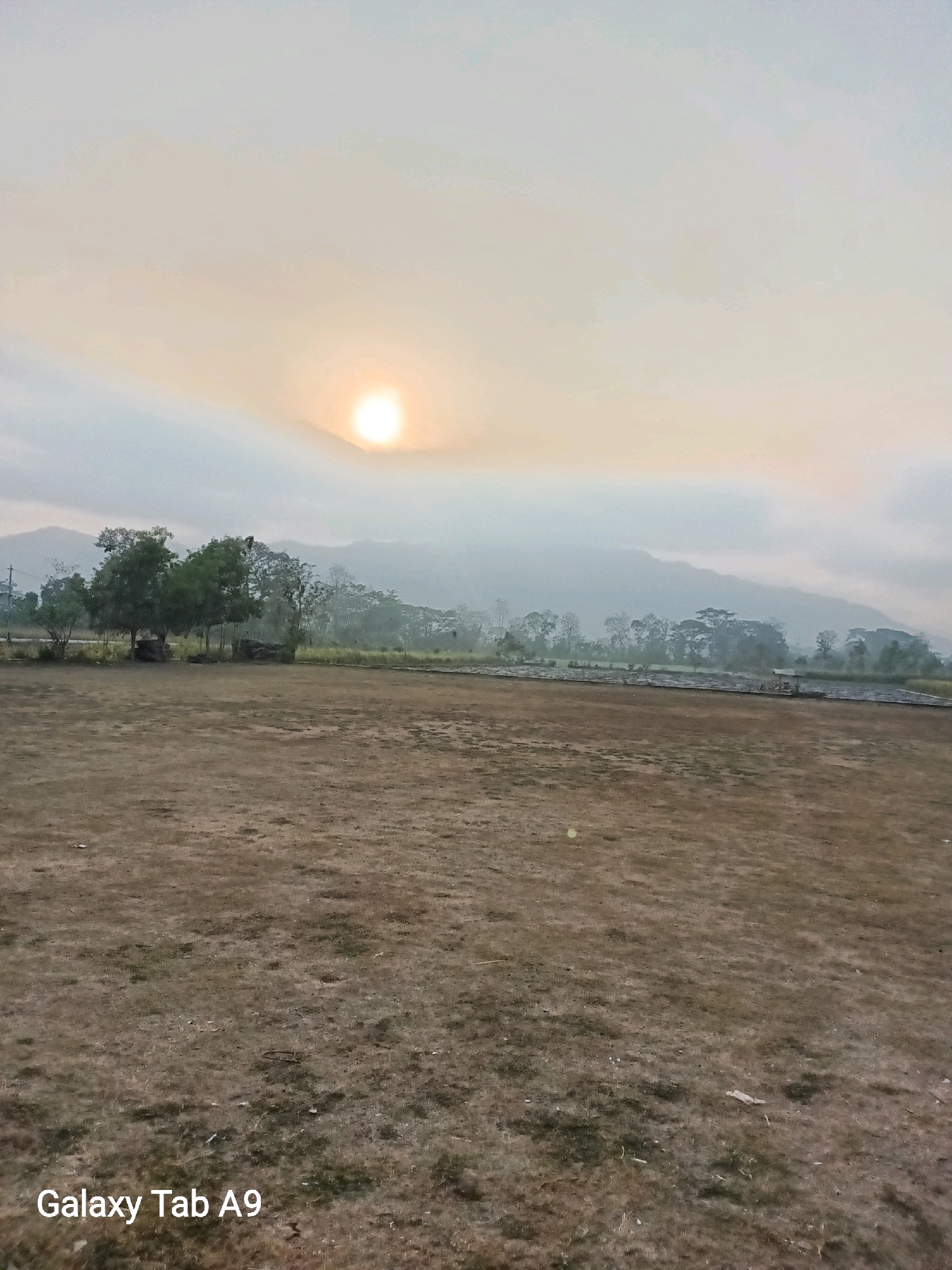 The image depicts an outdoor scene at sunrise in Bendorejo, Indonesia. The foreground is an open, dry, grassy field. In the middle ground, a line of trees with dense foliage stands against a backdrop of hazy mountains. The sun, a bright orb, is visible in the upper left portion of the sky, partially obscured by clouds. The overall atmosphere is serene and quiet, characteristic of early morning in a rural or semi-rural setting. The bottom left corner of the image displays the text "Galaxy Tab A9," indicating the device used to capture the photograph.