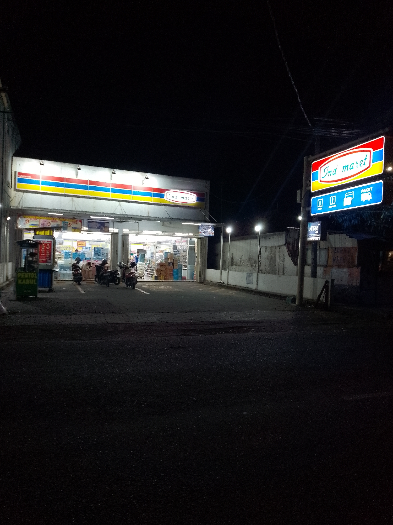 The image captures a night scene outside an Indomaret convenience store in Lamongan, Indonesia. The store's facade is illuminated by overhead lights, and its distinctive red, yellow, and blue striped awning is visible. A prominent Indomaret sign with the text "Indomaret" in red script on a yellow background is mounted on a pole to the right of the store. Below this, a blue sign displays icons for ATMs, credit cards, and packages.
In front of the store, several motorcycles are parked. One motorcycle has a rider wearing a white outfit. Another person is standing near the motorcycles, also in white attire. To the left of the store entrance, a green cart labeled "PENTOL" and "KABUL" stands near a blue bin. The area in front of the store appears to be paved, with a darker road surface in the foreground. The background consists of a textured wall and a power pole. The overall atmosphere is dim, with artificial lighting from the store and the signs illuminating the immediate surroundings.