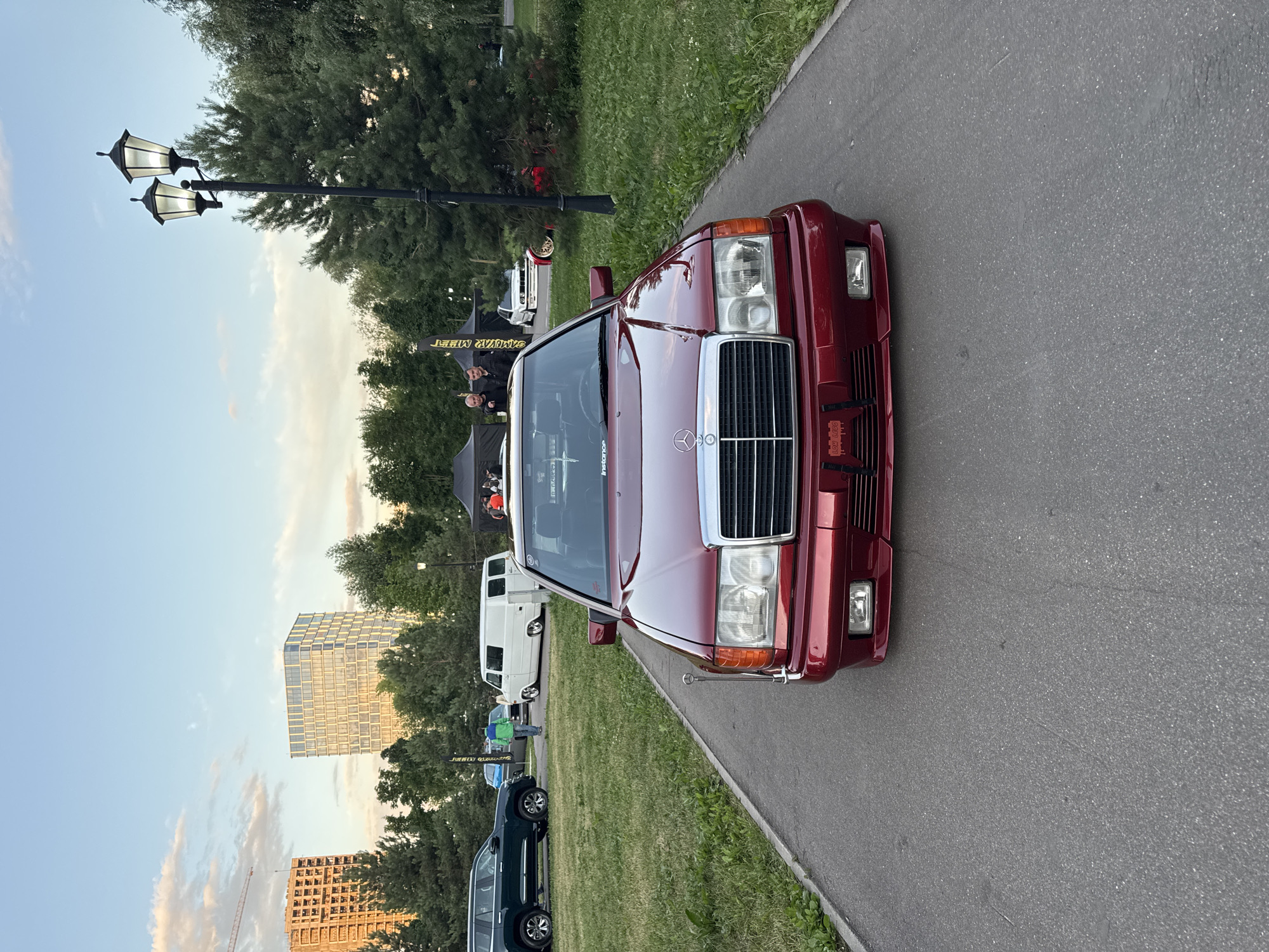 A maroon Mercedes-Benz W124 coupe is parked on an asphalt pathway in a park setting. The car is facing the camera, with its front grille, headlights, and bumper clearly visible. The car has a lowered stance, indicating it may be modified. In the background, several tents are set up, suggesting an outdoor event or gathering. There are trees and grass surrounding the pathway, typical of a park. In the far background, modern buildings and a construction crane are visible, indicating an urban environment. The sky is clear with some clouds, and the lighting suggests it is late afternoon or early evening. The scene is likely a car show or meet-up event in Moscow, Russia, given the presence of a Mercedes-Benz and the contemporary urban architecture.
