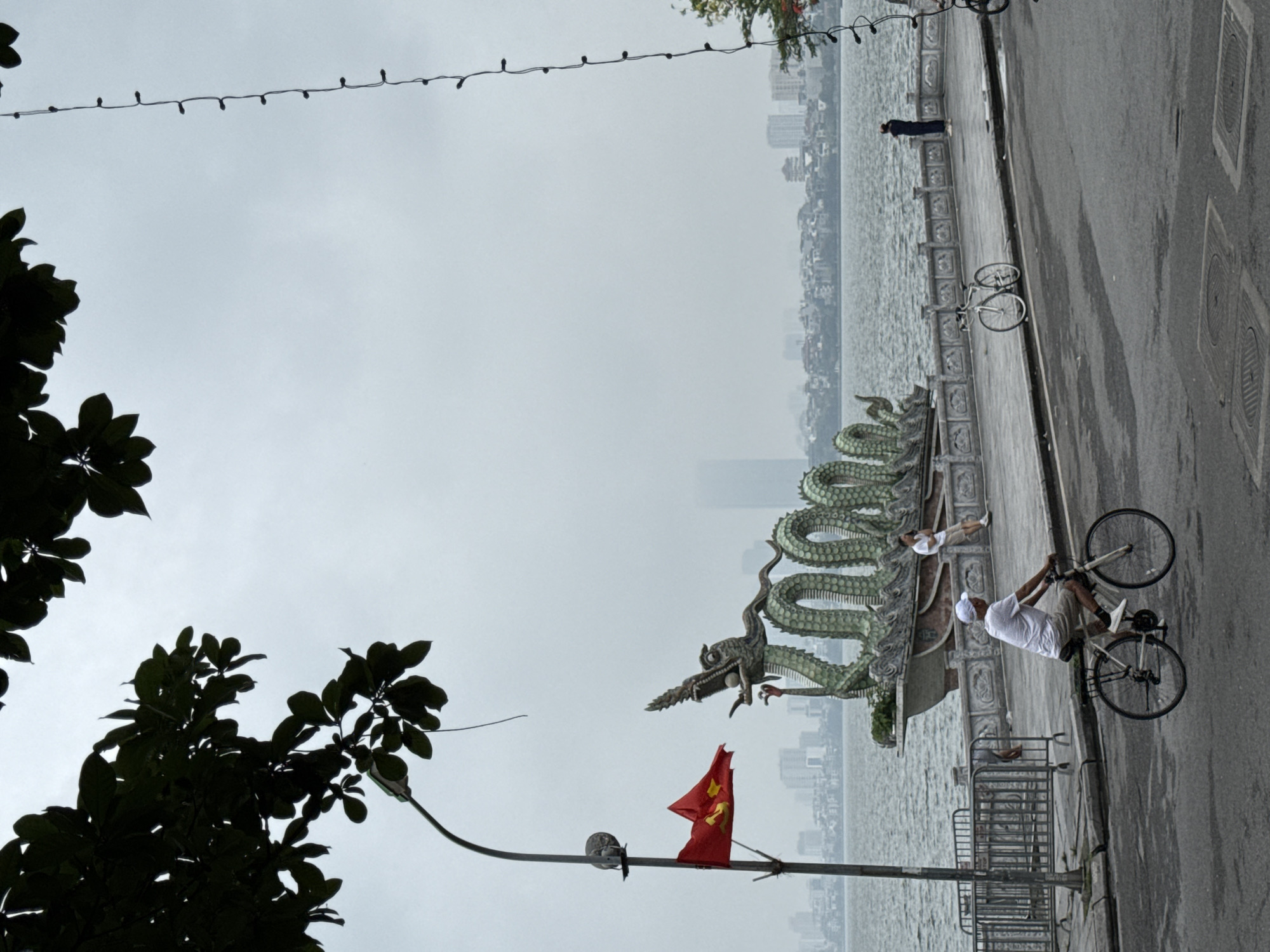 The image depicts a street scene in Hà Nội, Vietnam, specifically along the Hoàn Kiếm Lake. A large, sculpted dragon monument stands along the lake's edge. A Vietnamese flag is attached to a street lamp. A man rides a bicycle along the pavement, and another person rests on the dragon monument. Buildings are visible across the lake in the distance. The sky is overcast.